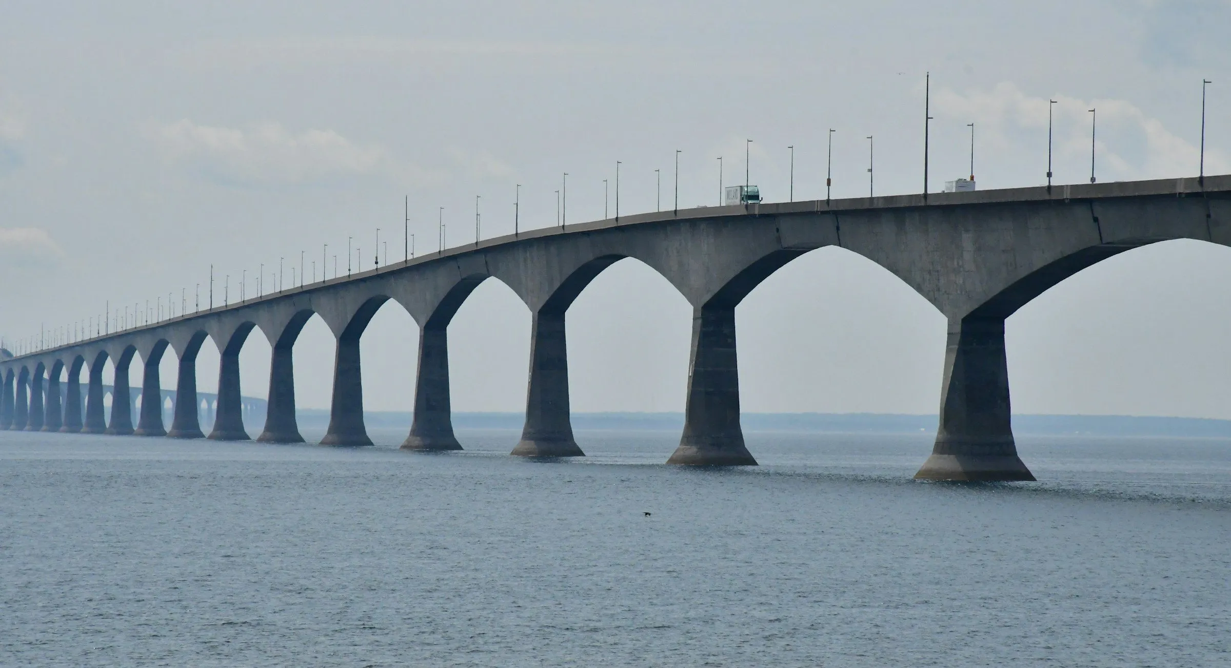 The Confederation Bridge curving away across the Northumberland Strait on tall concrete piers, under a hazy sky.