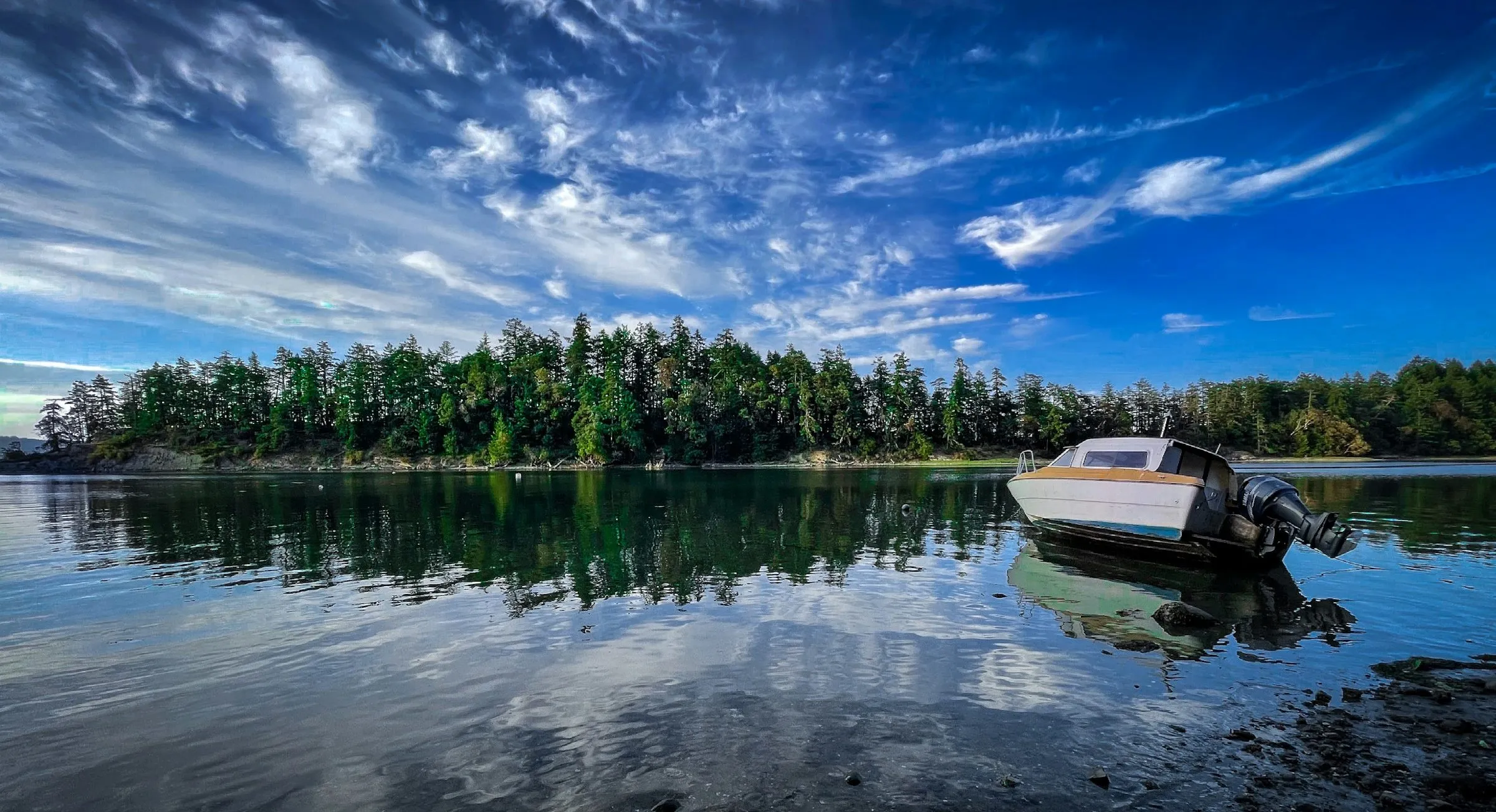 A small motorboat rests in still shallow water off Salt Spring Island, with a forested shore reflected under streaks of high cloud.