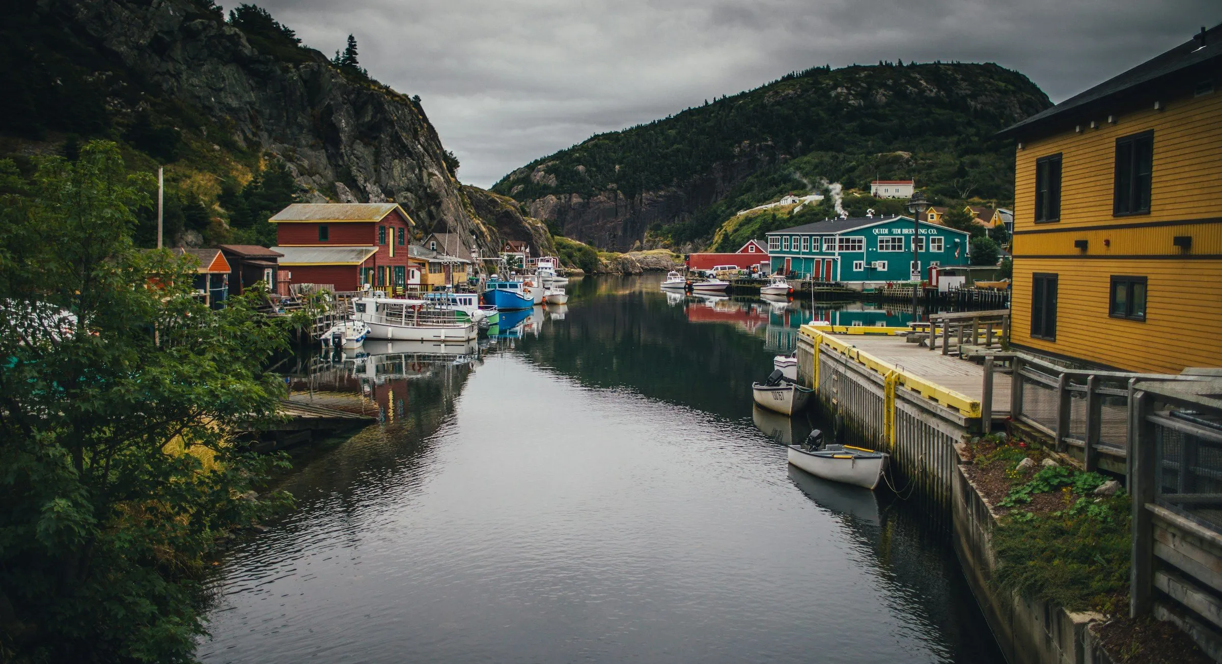 A narrow Newfoundland harbour lined with fishing stages, small boats and painted wooden buildings under a dark sky.