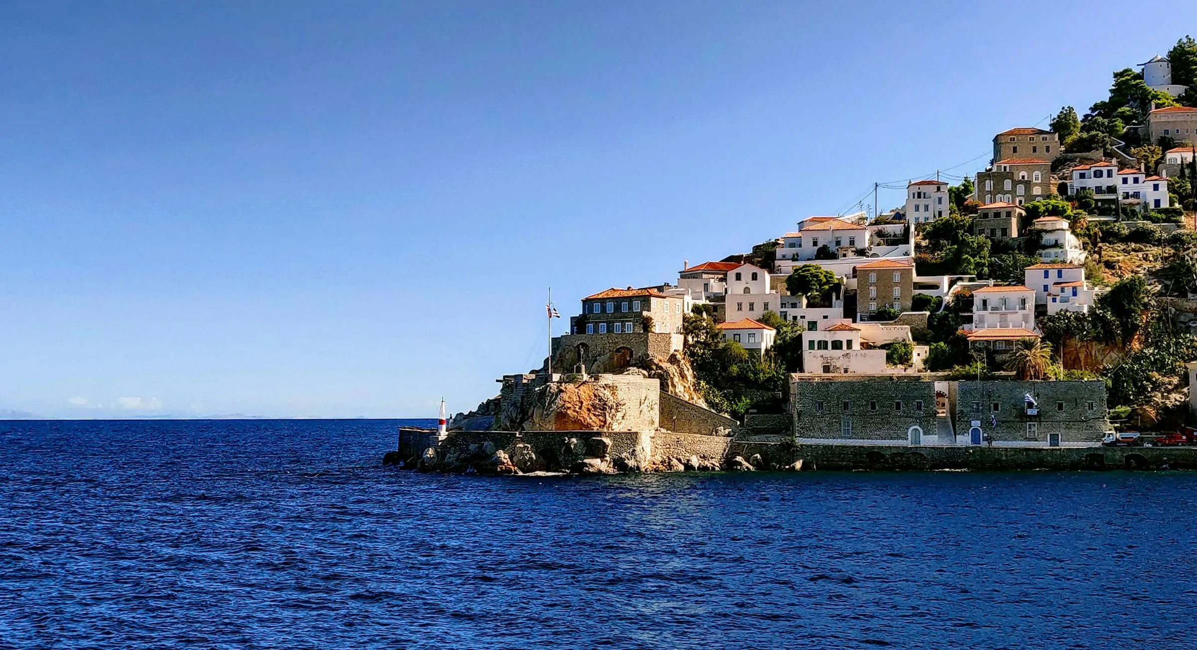 Stone and whitewashed houses with terracotta roofs climb a rocky headland above deep blue sea on Hydra.