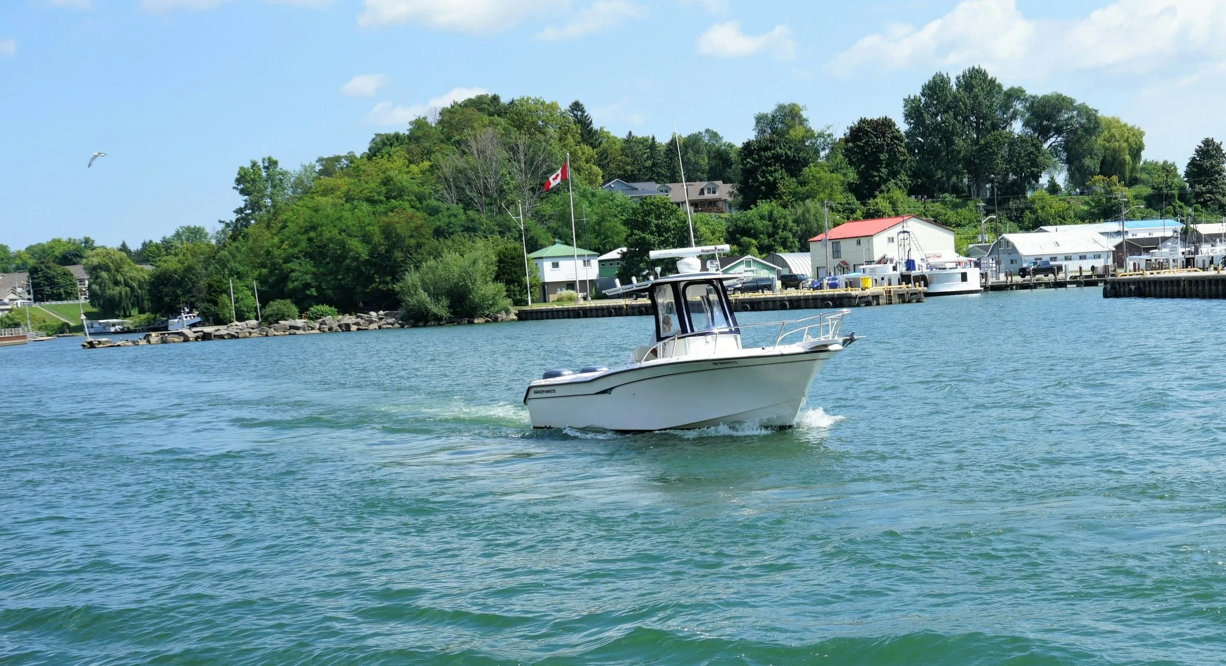 A white motorboat crosses the harbour mouth at Port Dover, with a Canadian flag, boathouses and trees along the shore behind it.