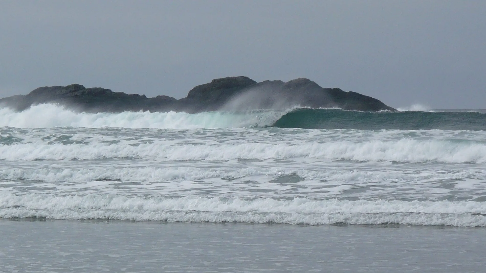 Lines of large grey waves breaking towards Chesterman Beach, with spray blowing off their tops and a rocky island behind.