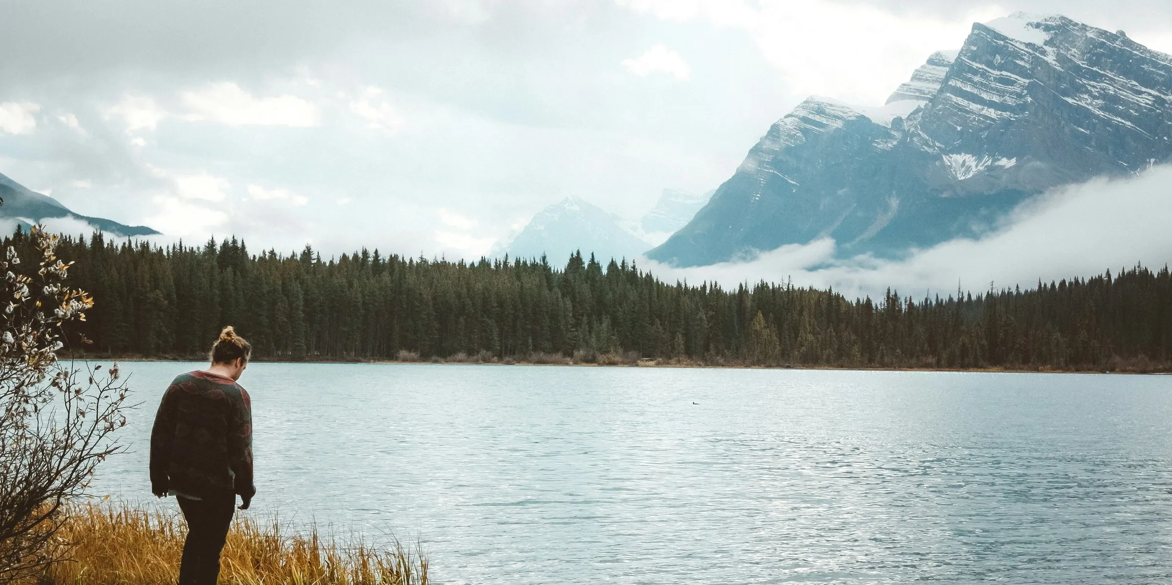A mountain lake under snow-dusted peaks and low cloud, with dark forest along the far shore.
