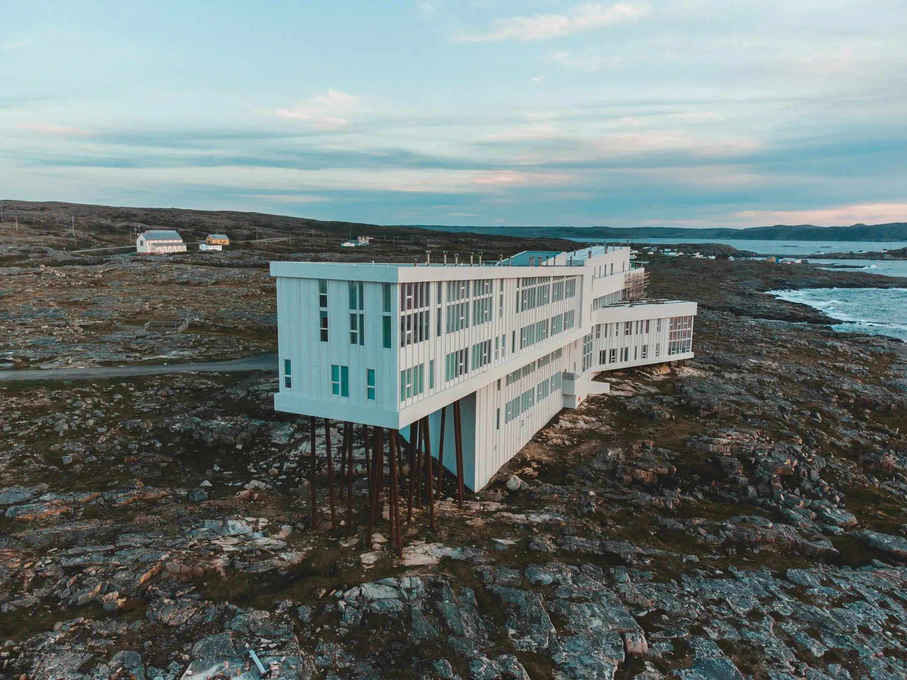The Fogo Island Inn seen from the air, an X-shaped white building raised on legs above bare rock, with the sea behind.