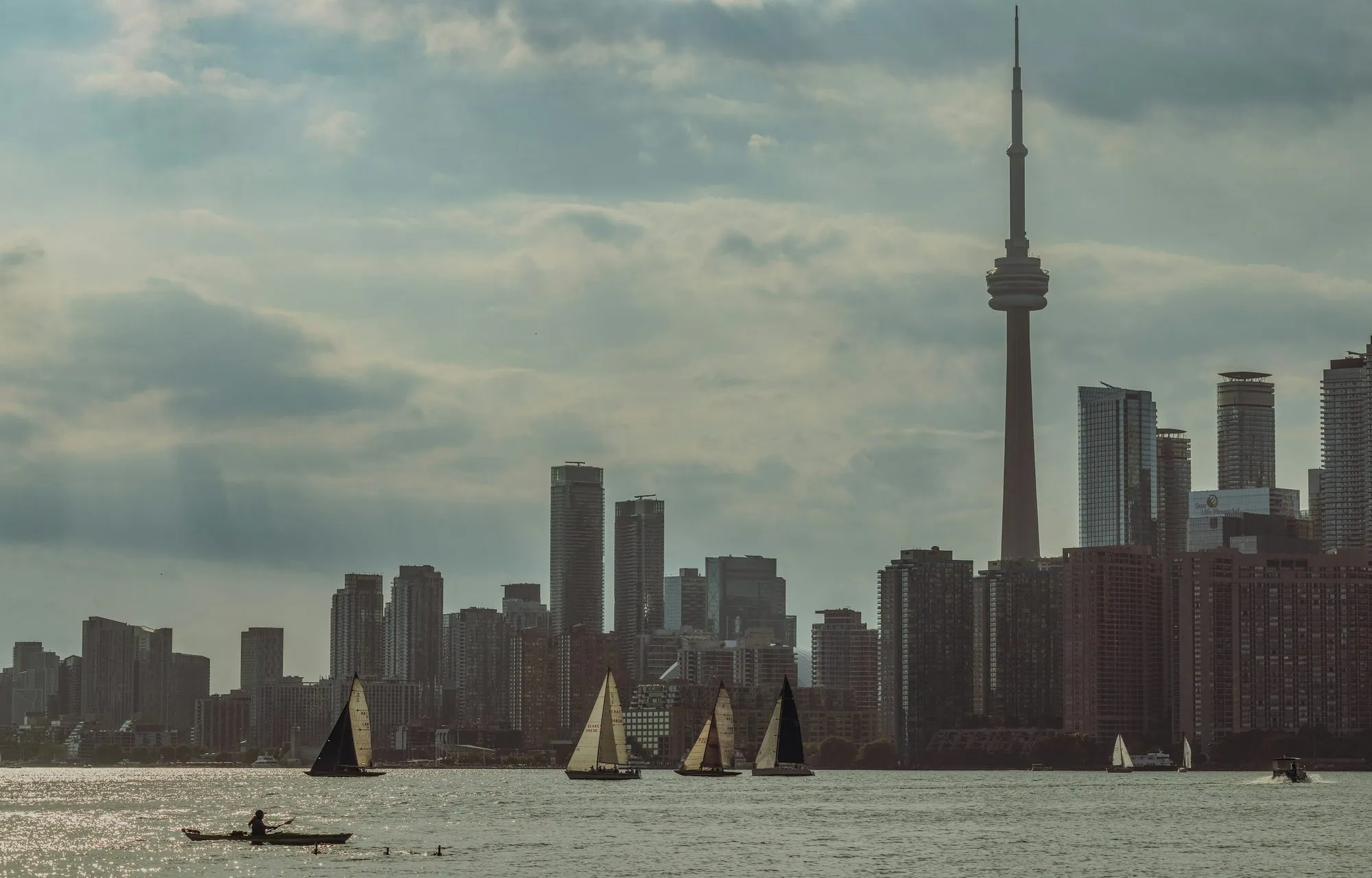 Sailboats on Lake Ontario in front of the Toronto skyline and the CN Tower, under a cloudy sky.