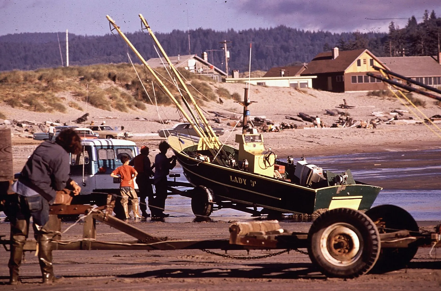 A dory on a trailer on the beach with its crew standing around it, and houses on the dune behind.