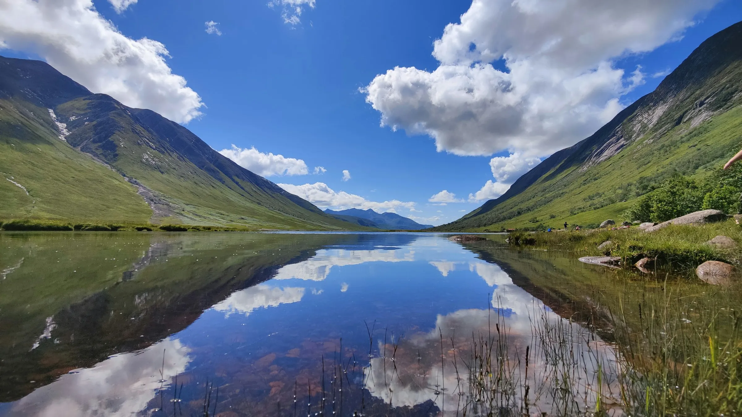 Mountains and clouds reflected in still water in Glen Etive under a blue sky.