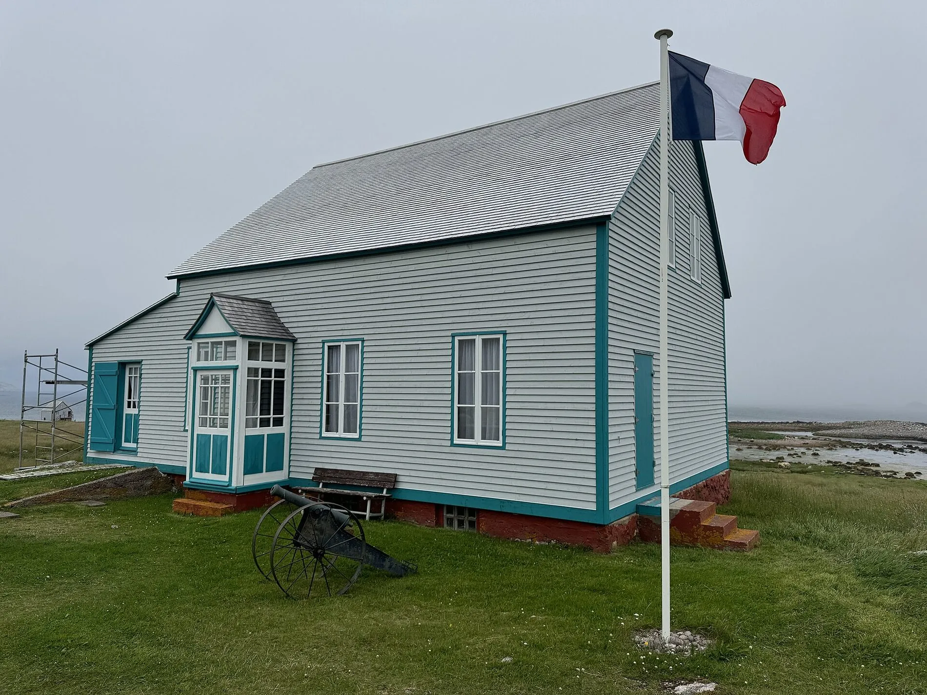 A grey wooden house on Île aux Marins with a French tricolour flying from a pole beside it and an old anchor on the grass.