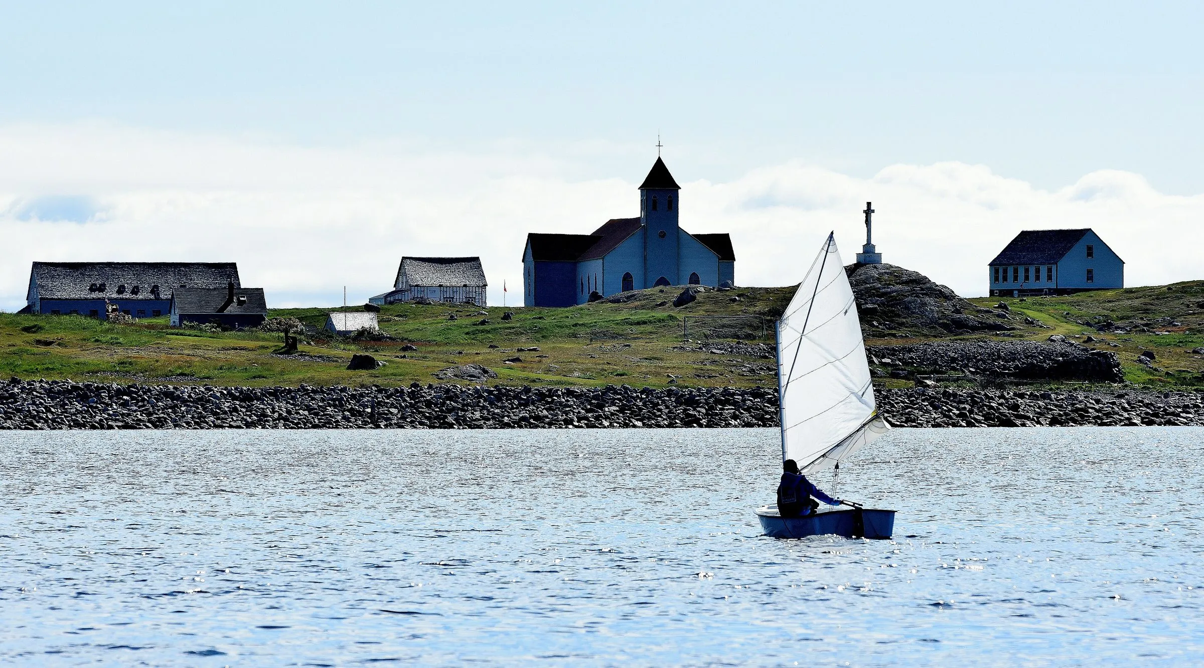 A small boat under a white sail passing Île aux Marins, with its church and a few houses on the low shore.