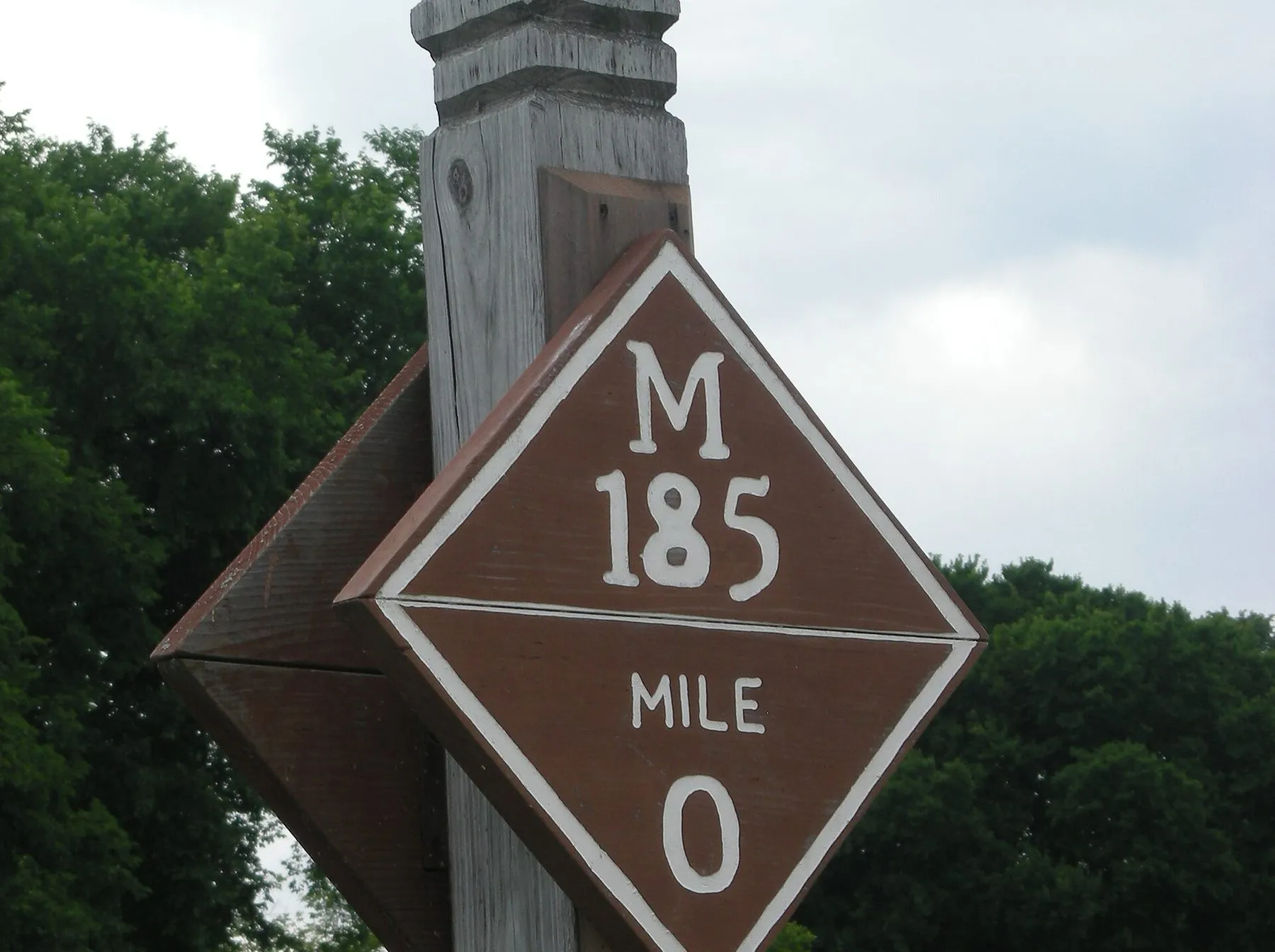 A brown diamond-shaped wooden sign on a post reading M 185, Mile 0.