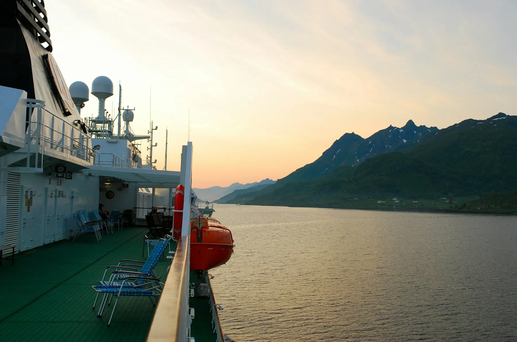 The side deck of a Hurtigruten ship with an orange lifeboat and deck chairs, looking across calm water to mountains under a glowing sky.