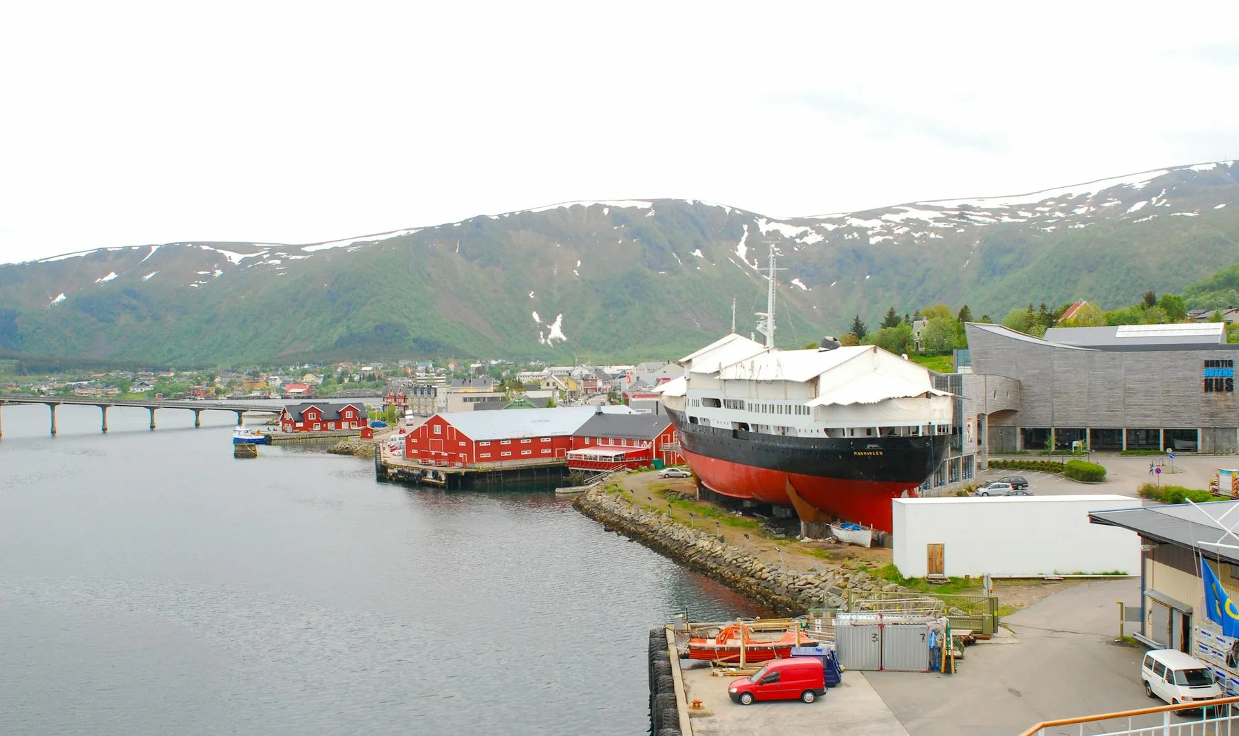 The harbour at Stokmarknes, with a black, white and red ship standing on land beside the museum buildings and green mountains behind.