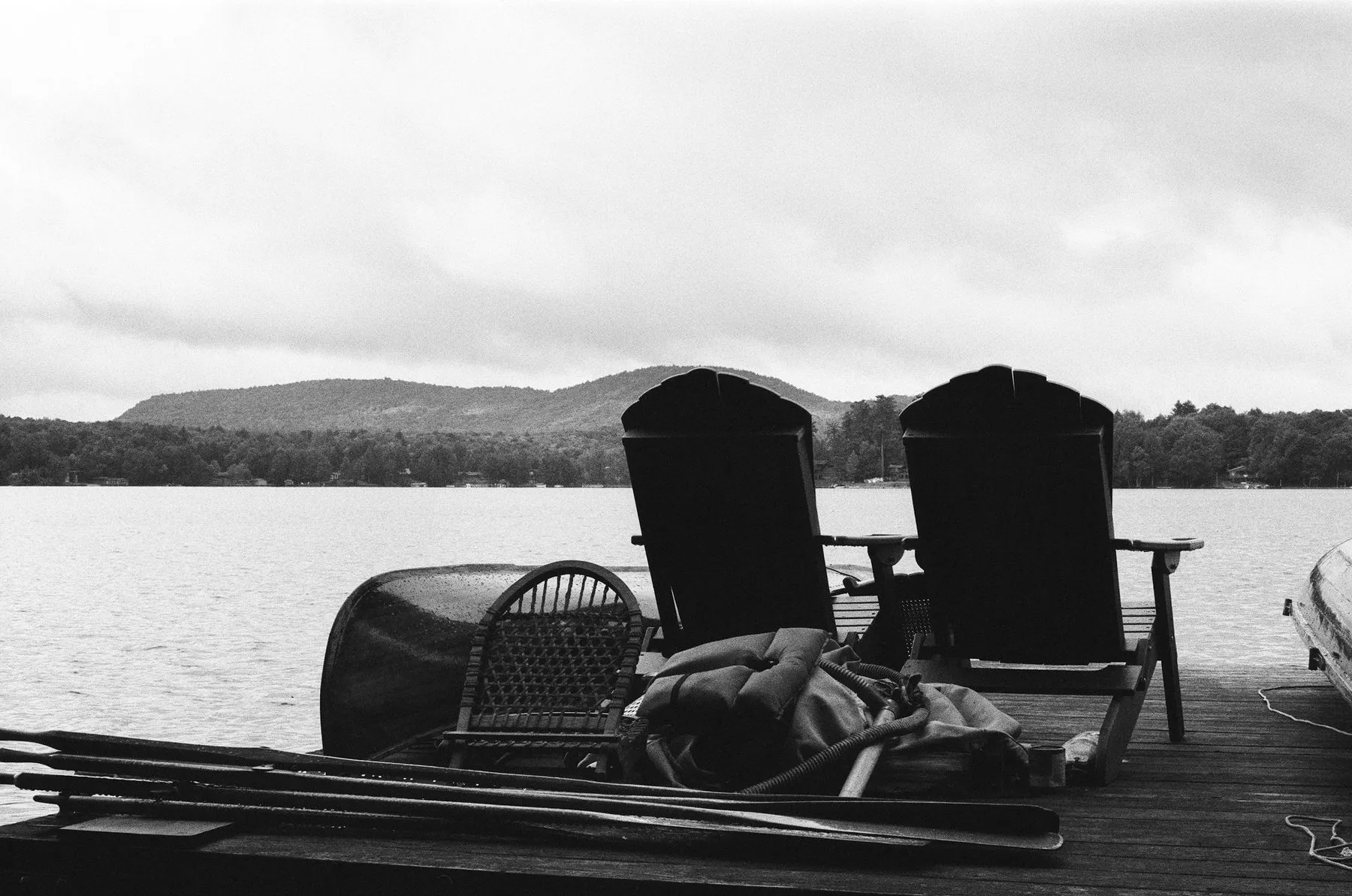 A black and white photo of two dark Adirondack chairs on a dock, facing a lake and low mountains.