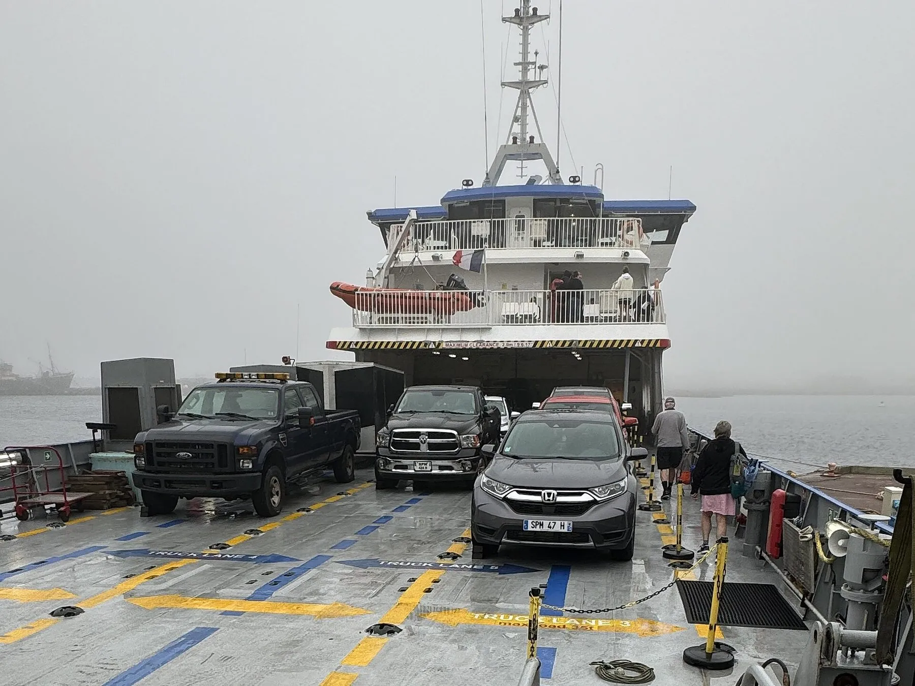 Pickup trucks and cars parked on the open deck of the ferry in fog, below the wheelhouse.