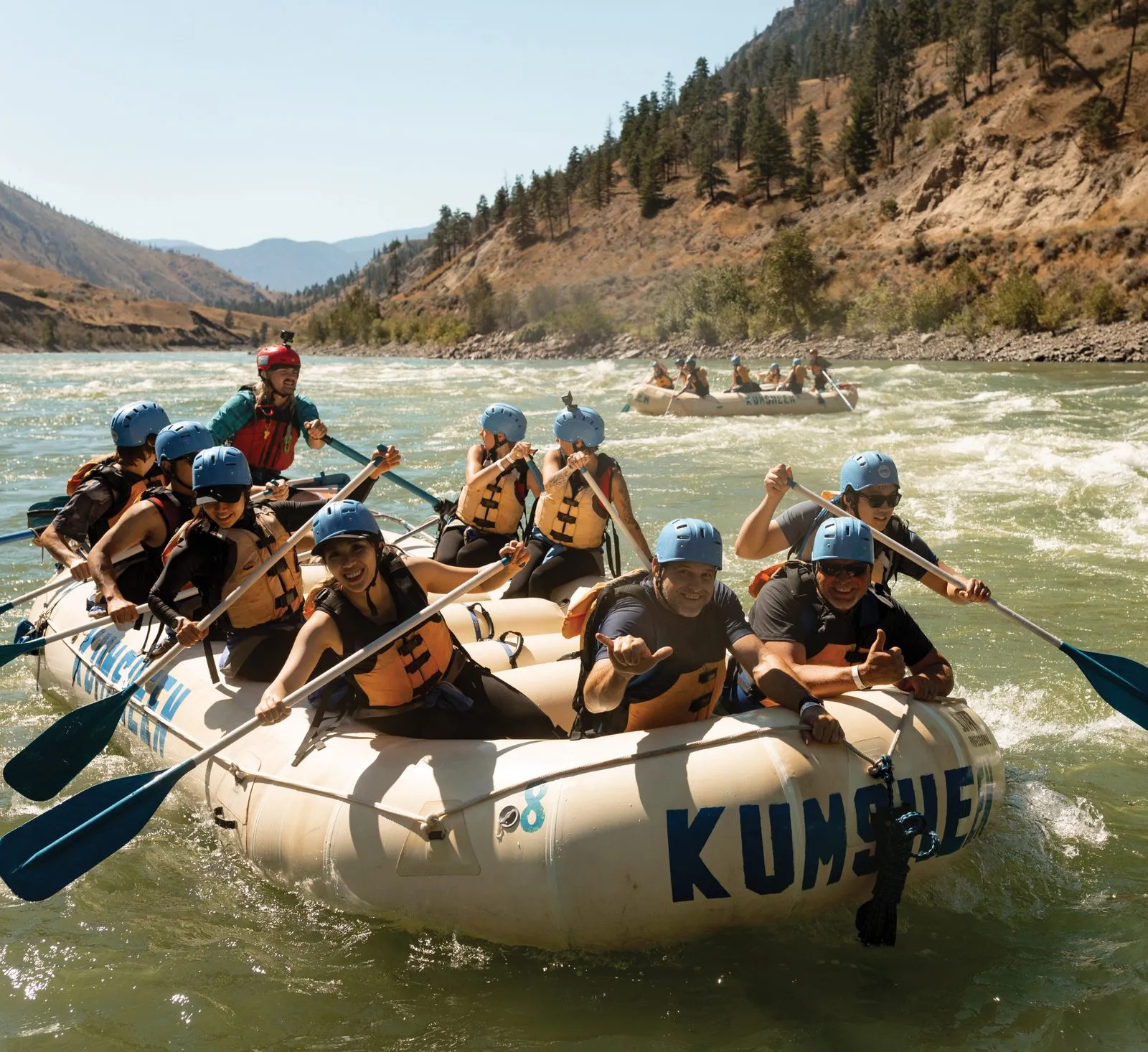 Eight people in helmets and orange life jackets paddle a raft marked Kumsheen, grinning at the camera, with dry hills behind.
