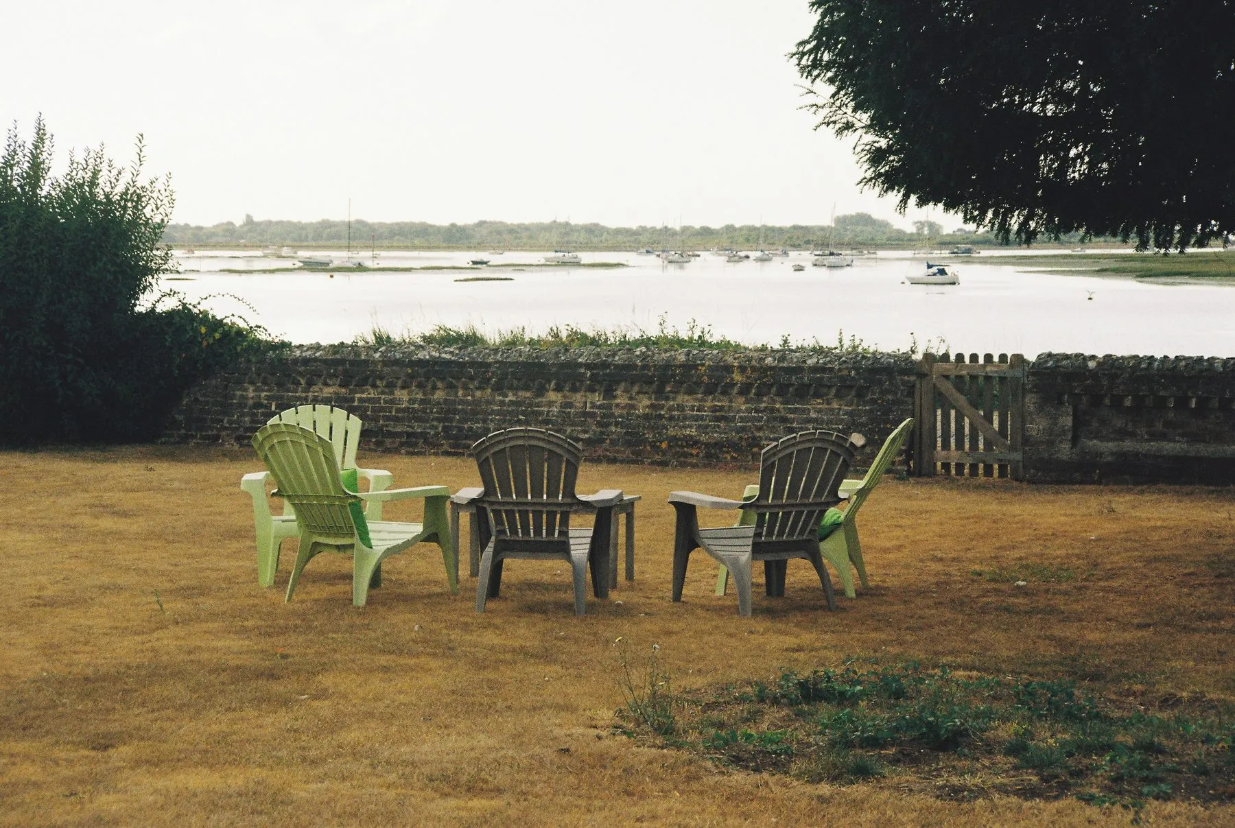 Four moulded plastic chairs in green and grey on a dry lawn, facing a bay with boats at anchor.