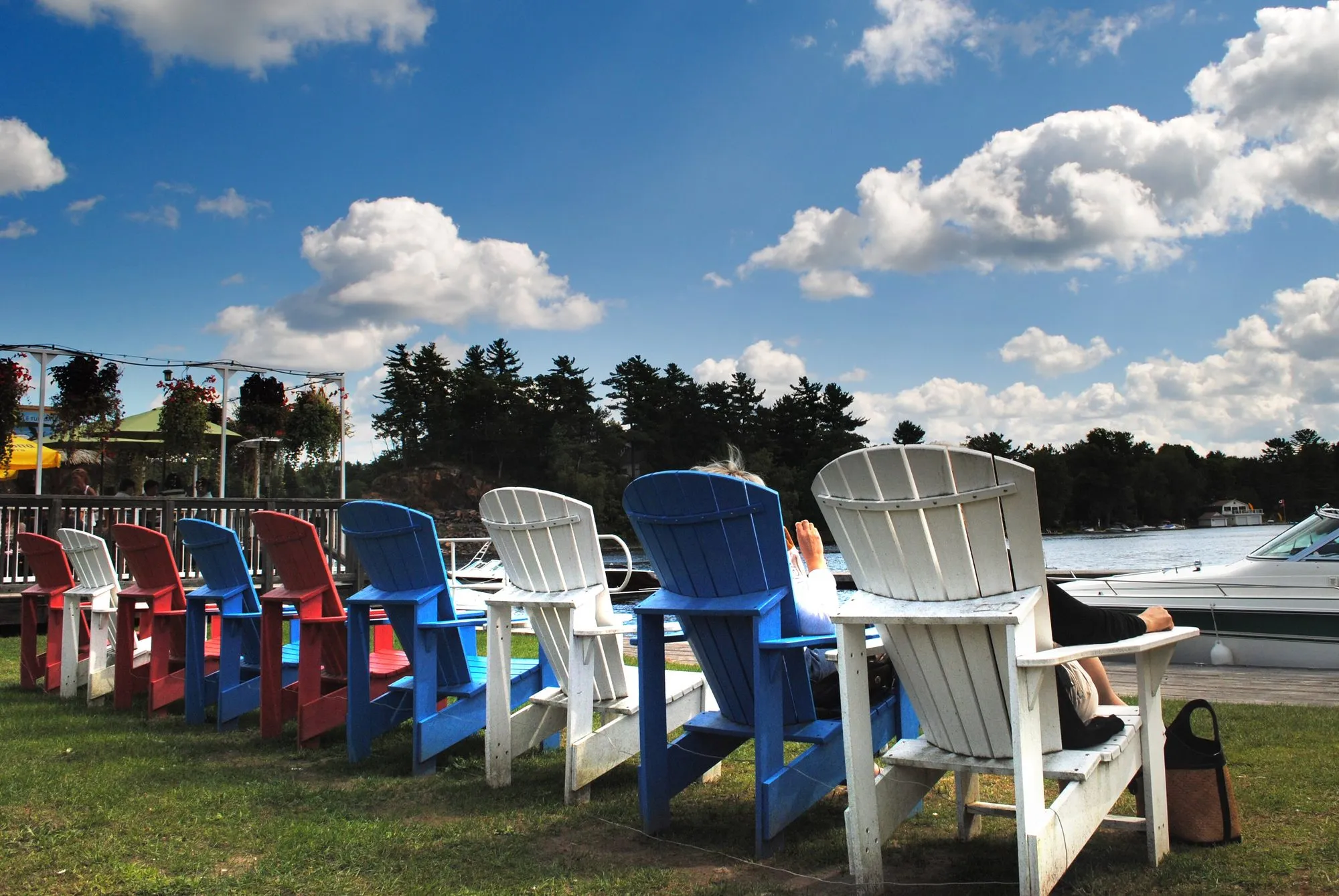 A long row of red, blue and white Muskoka chairs on a lawn beside the water at Port Carling, under a blue sky with white clouds.
