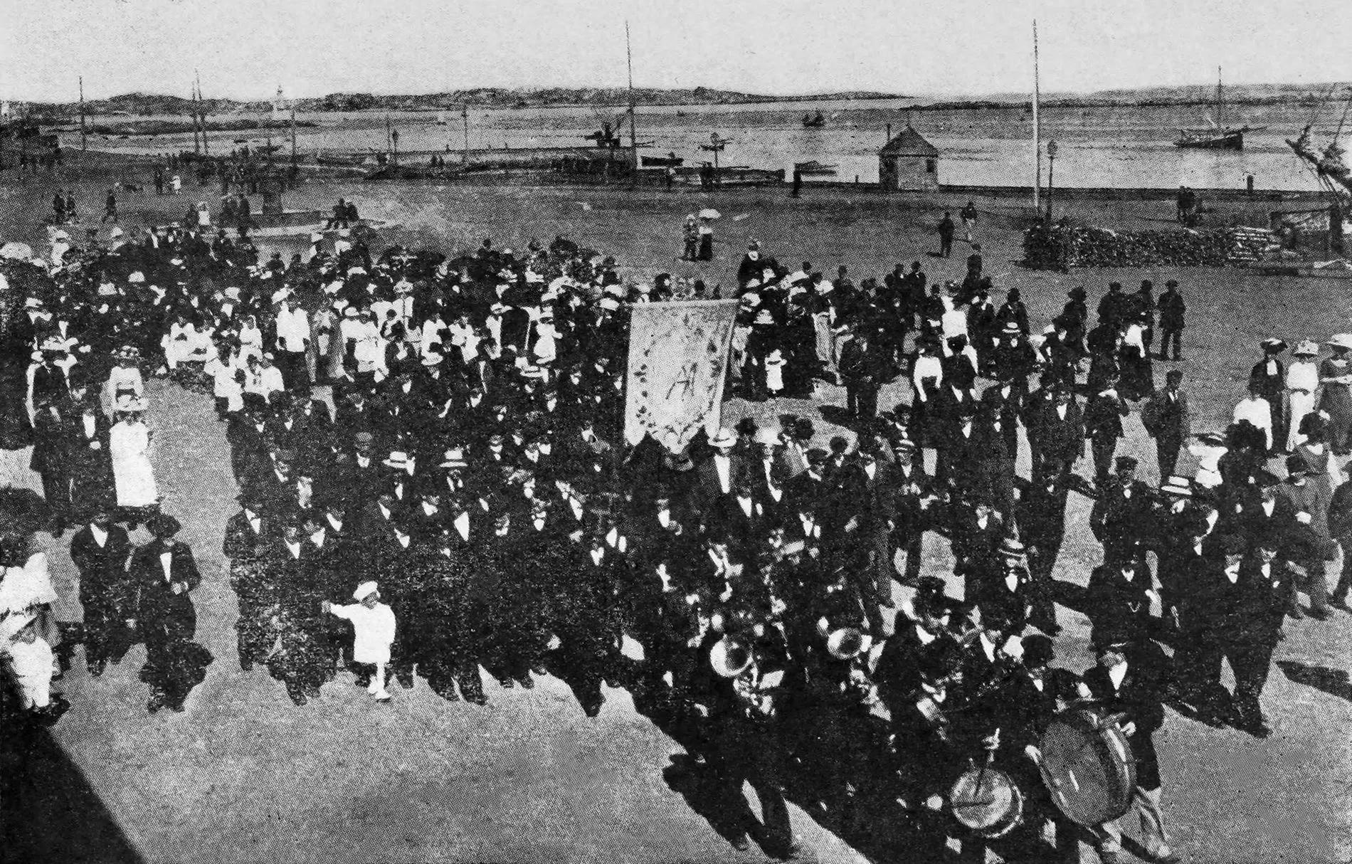 A black and white photograph of a large crowd in dark clothes gathered on the quay at Saint-Pierre, with boats in the harbour behind.