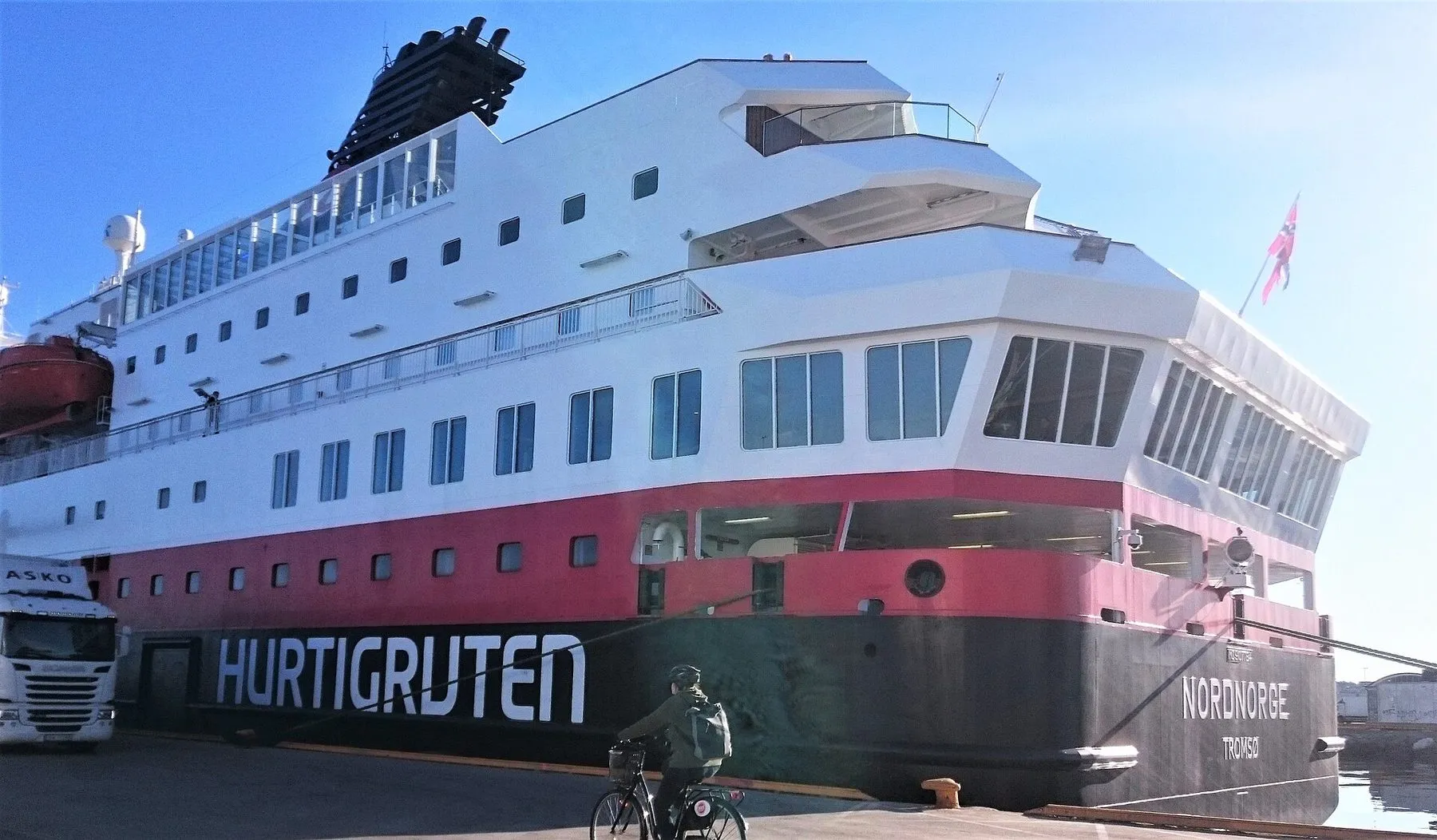 The stern of MS Nordnorge at a quay in Trondheim, with Hurtigruten written along the hull, a lorry alongside and a cyclist riding past.