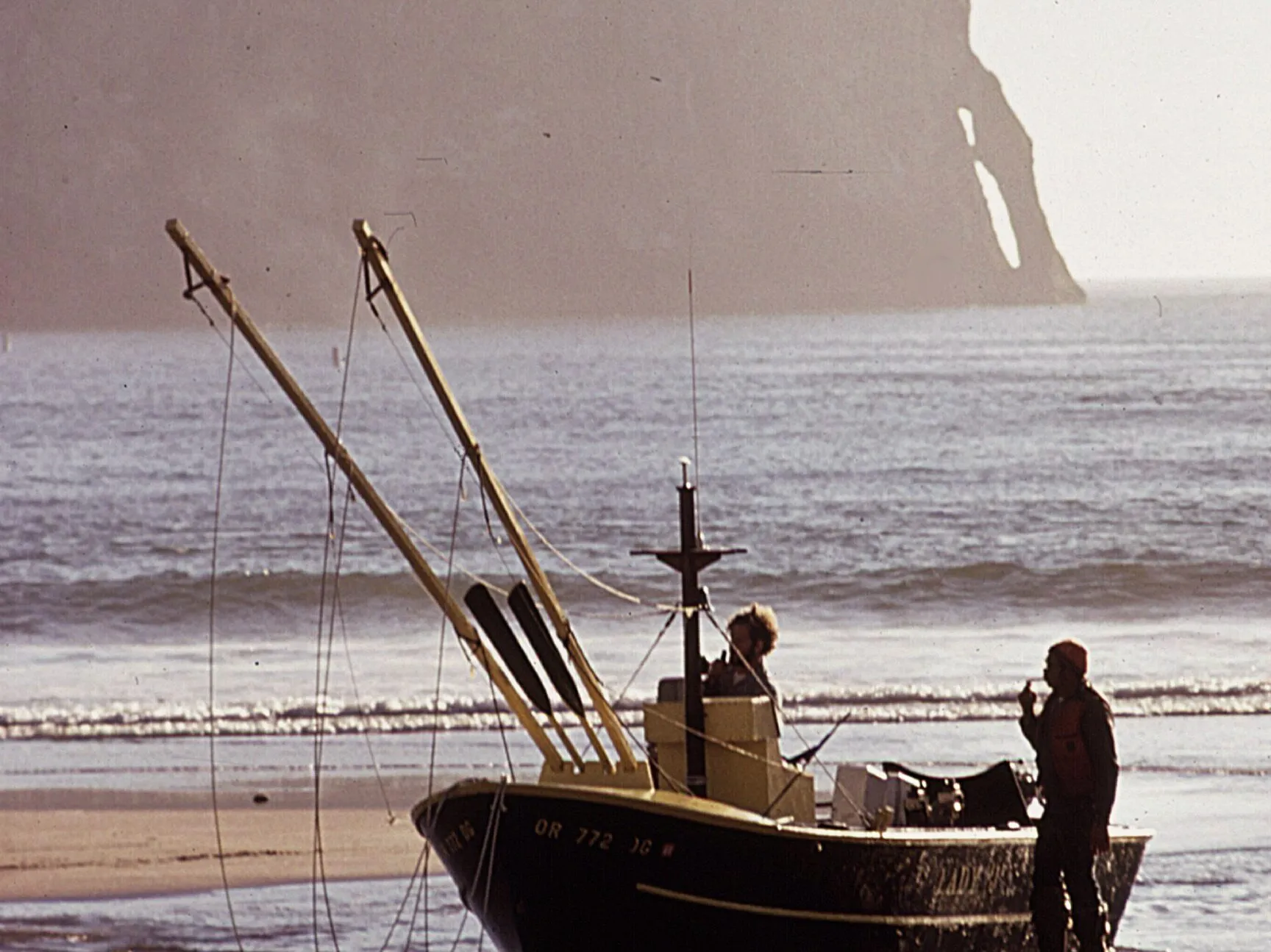 A dory sitting in the shallows with two fishermen beside it, and the foot of Haystack Rock in the haze behind.