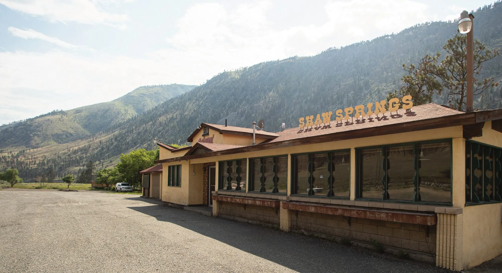 A low yellow roadside restaurant with a Shaw Springs sign on its roof, under dry forested hills.