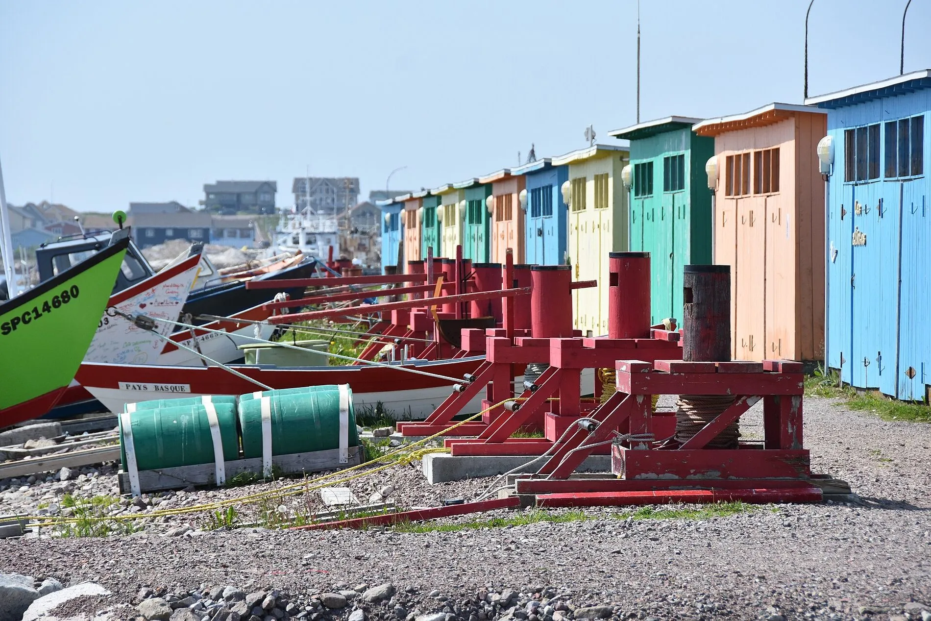 A row of narrow fishing sheds painted blue, orange, yellow and green, with red capstans and boats on the shingle in front.