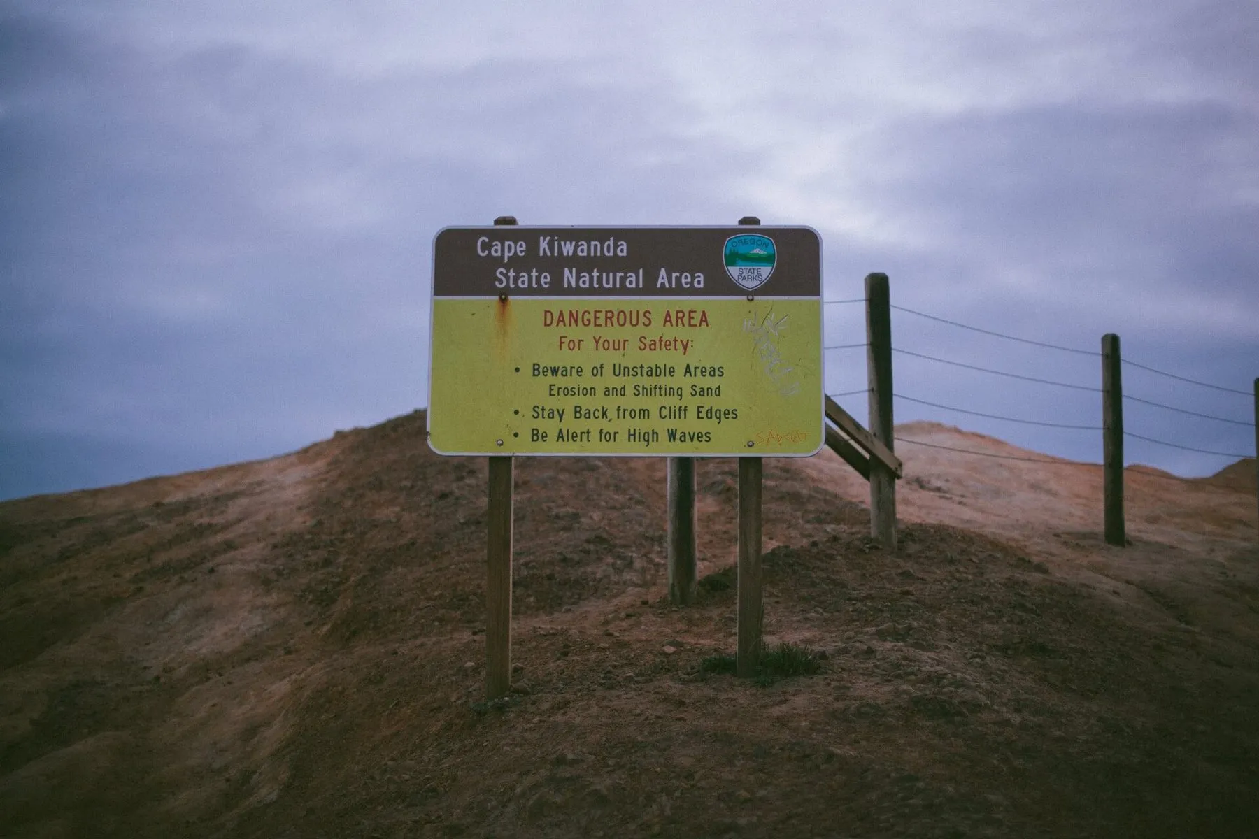 A yellow warning sign at Cape Kiwanda State Natural Area reading Dangerous Area, in front of a wire fence on a sandy bluff.