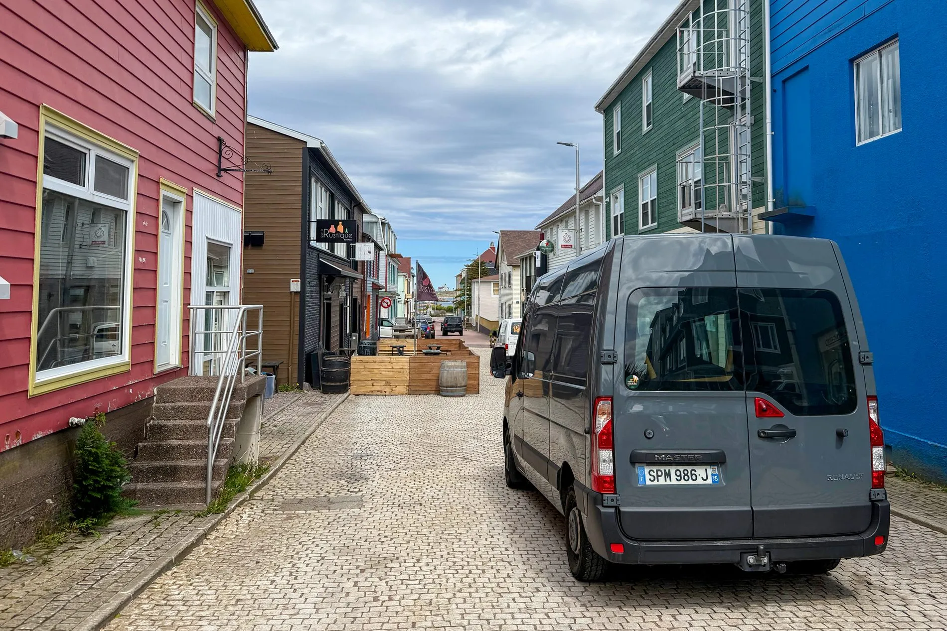 A narrow paved street between red, green and blue wooden buildings, with a grey van parked on the cobbles.