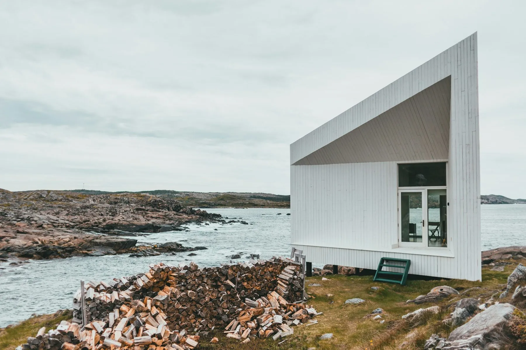 A small white wedge-shaped studio with a glass door, standing alone on a rocky shore beside a stack of firewood.