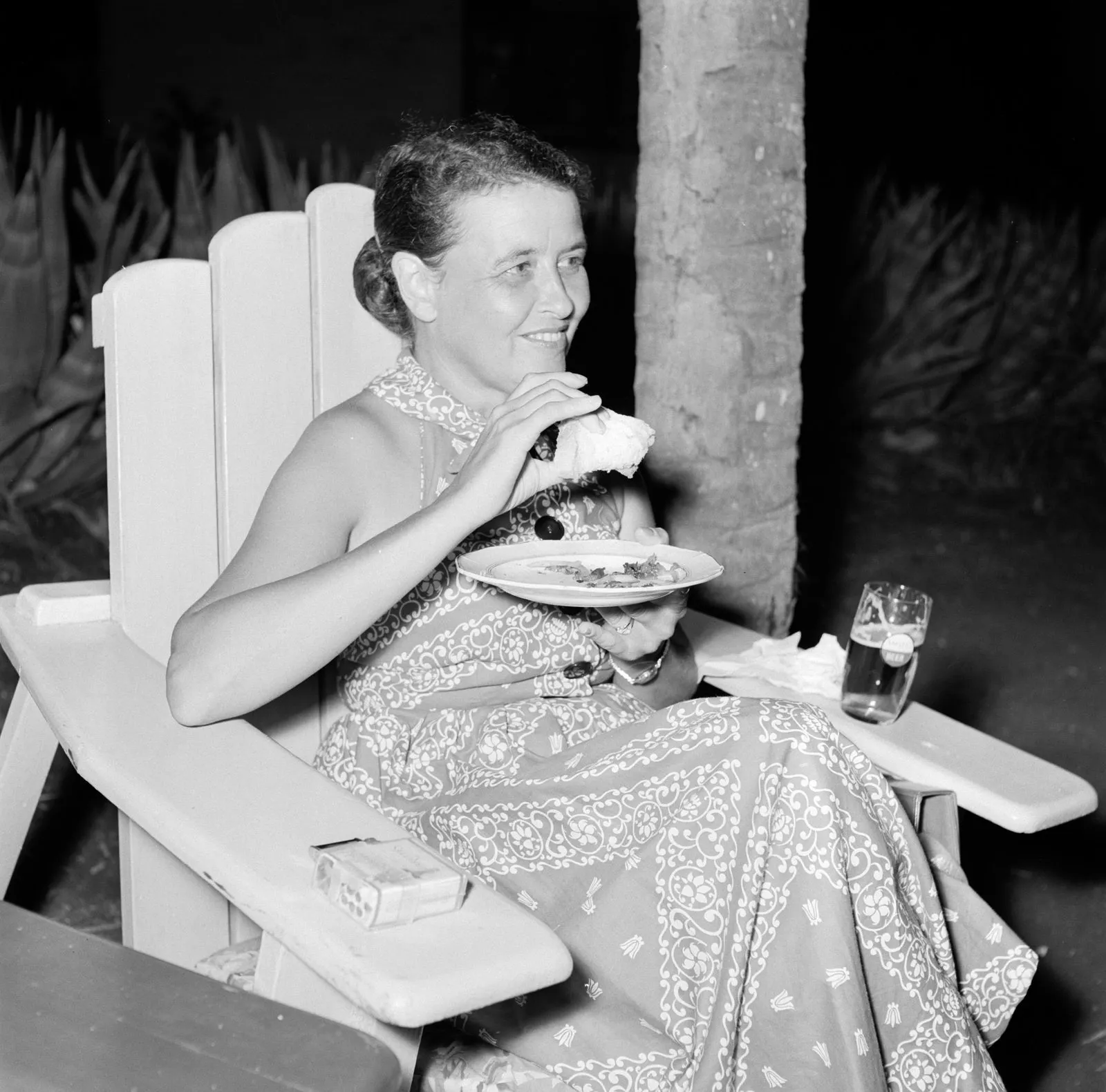 A black and white photo from 1955 of a woman in a patterned dress eating from a plate while sitting in a white Adirondack chair, with a drink on the wide arm.