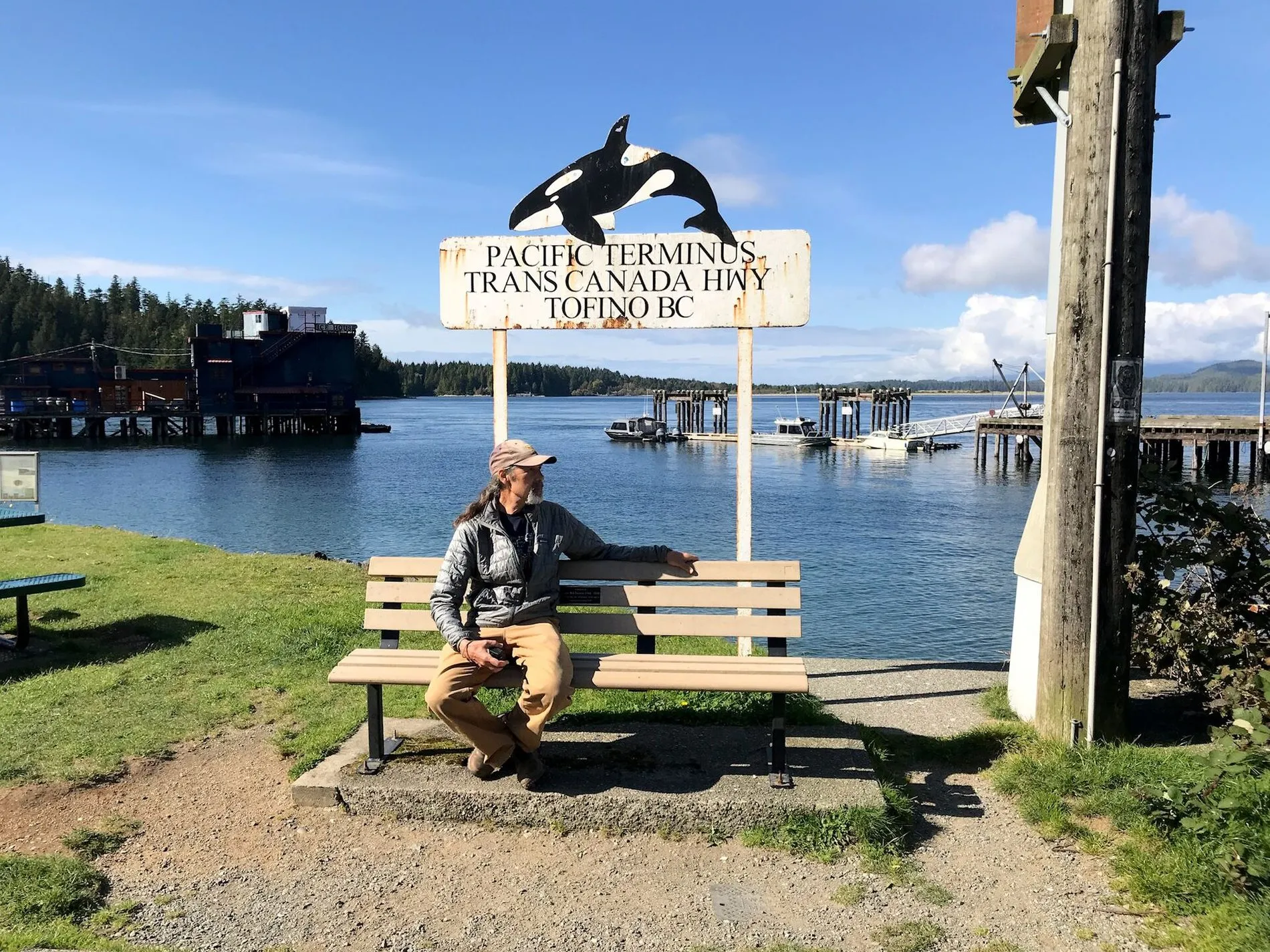 A man sitting on a bench by the harbour under a sign topped with an orca that reads Pacific Terminus, Trans Canada Hwy, Tofino BC.