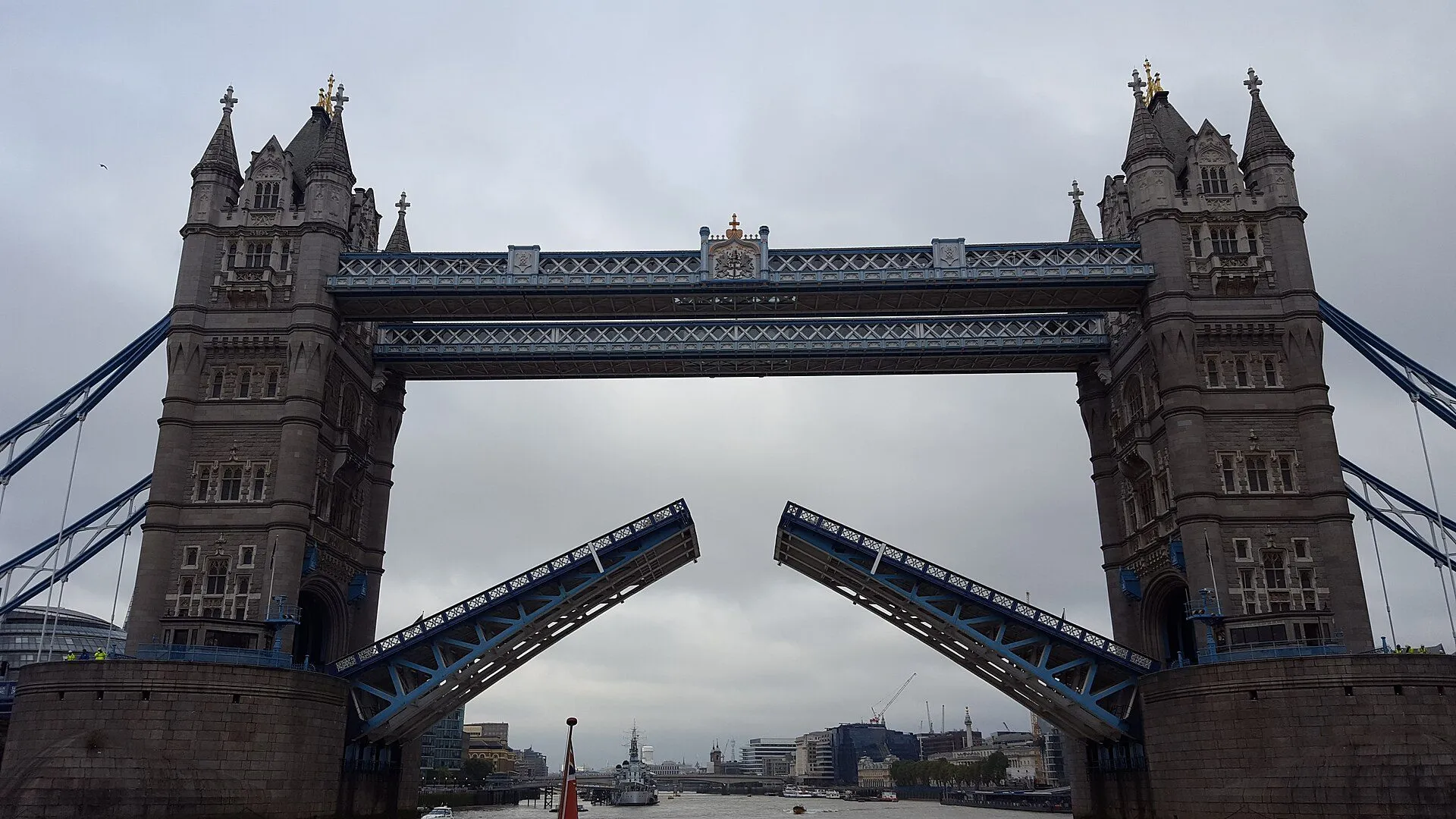 Tower Bridge in London with both halves of its roadway raised, seen from the river on a grey day.