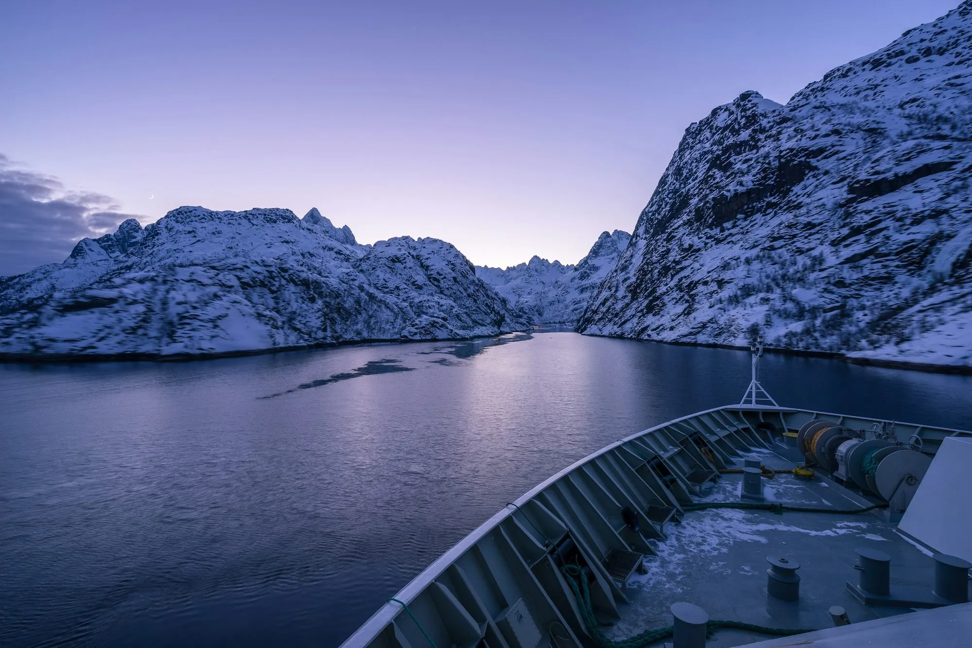 The bow of a ship entering the narrow Trollfjord between snow-covered mountains in blue evening light.
