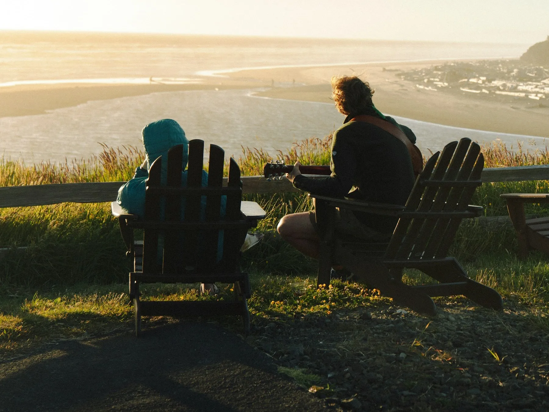 Two people relax in Adirondack chairs overlooking a beach and the ocean at sunset.