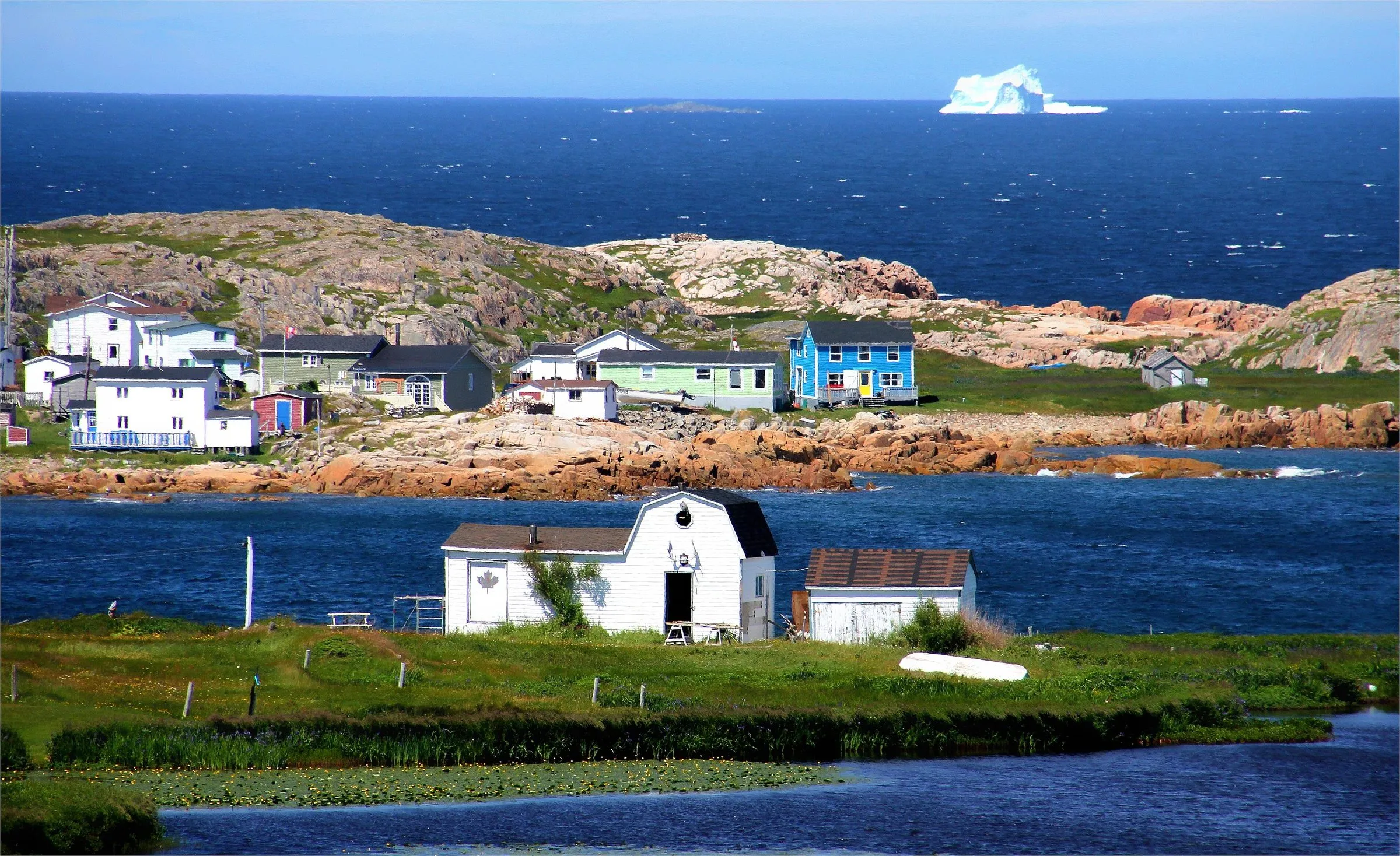 Brightly painted wooden houses on a rocky shore on Fogo Island, with an iceberg on the horizon.