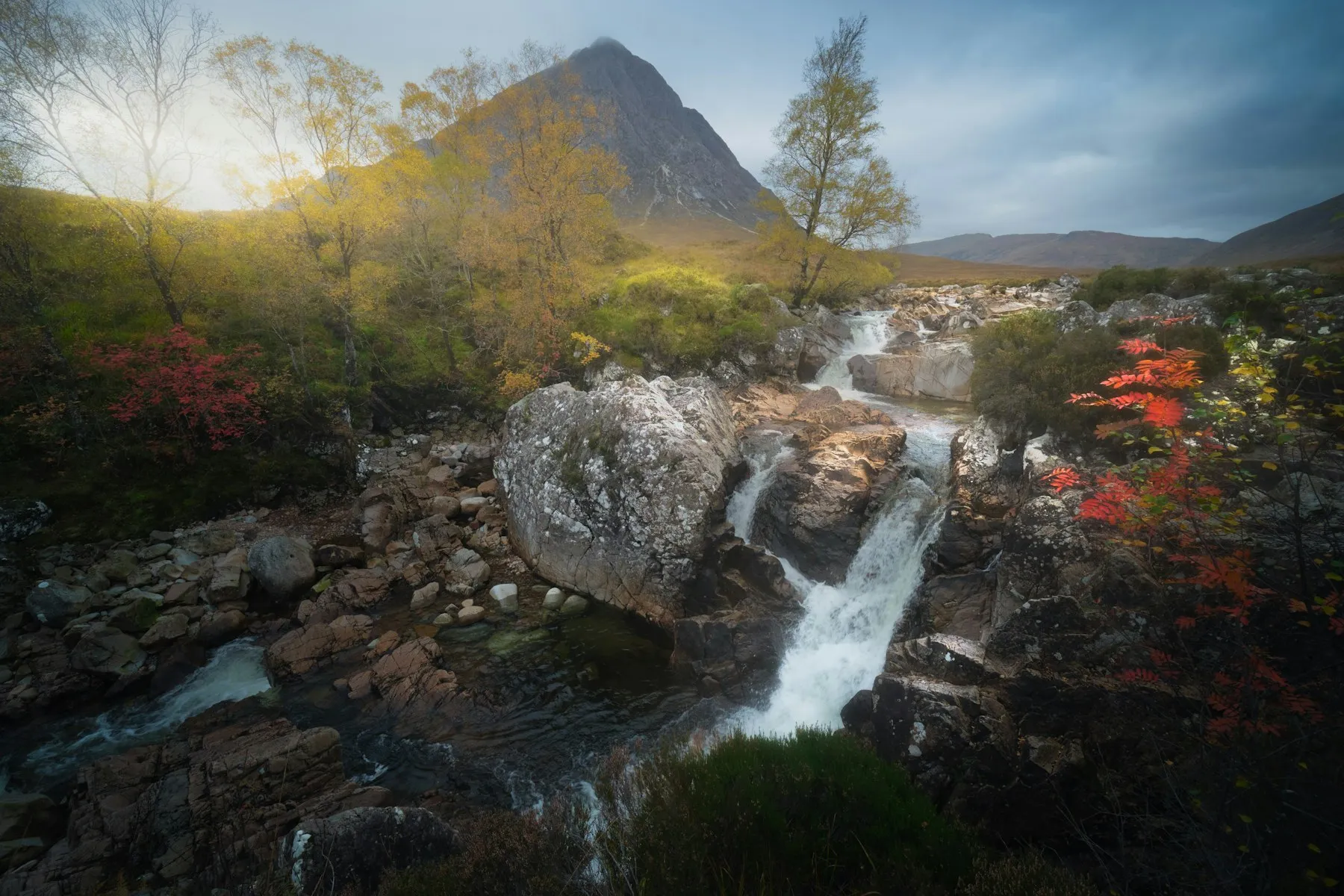 A waterfall among autumn trees, with the peak of Buachaille Etive Mòr behind it.