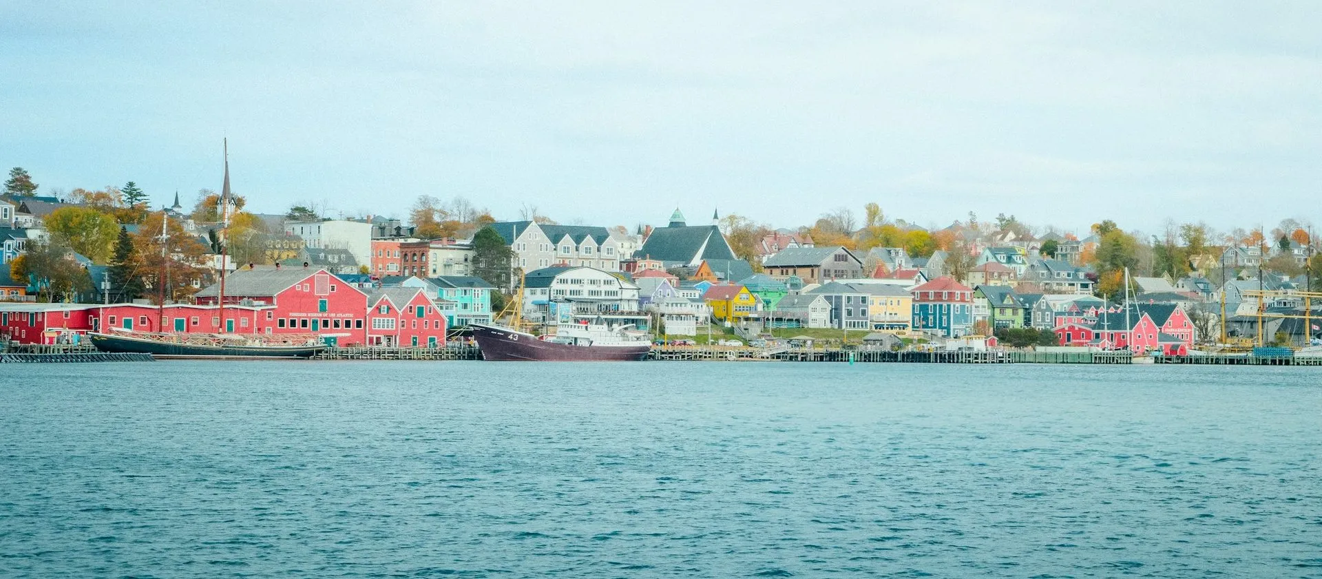 Lunenburg’s waterfront seen from across the harbour, a long row of red, yellow and blue buildings under a pale sky.