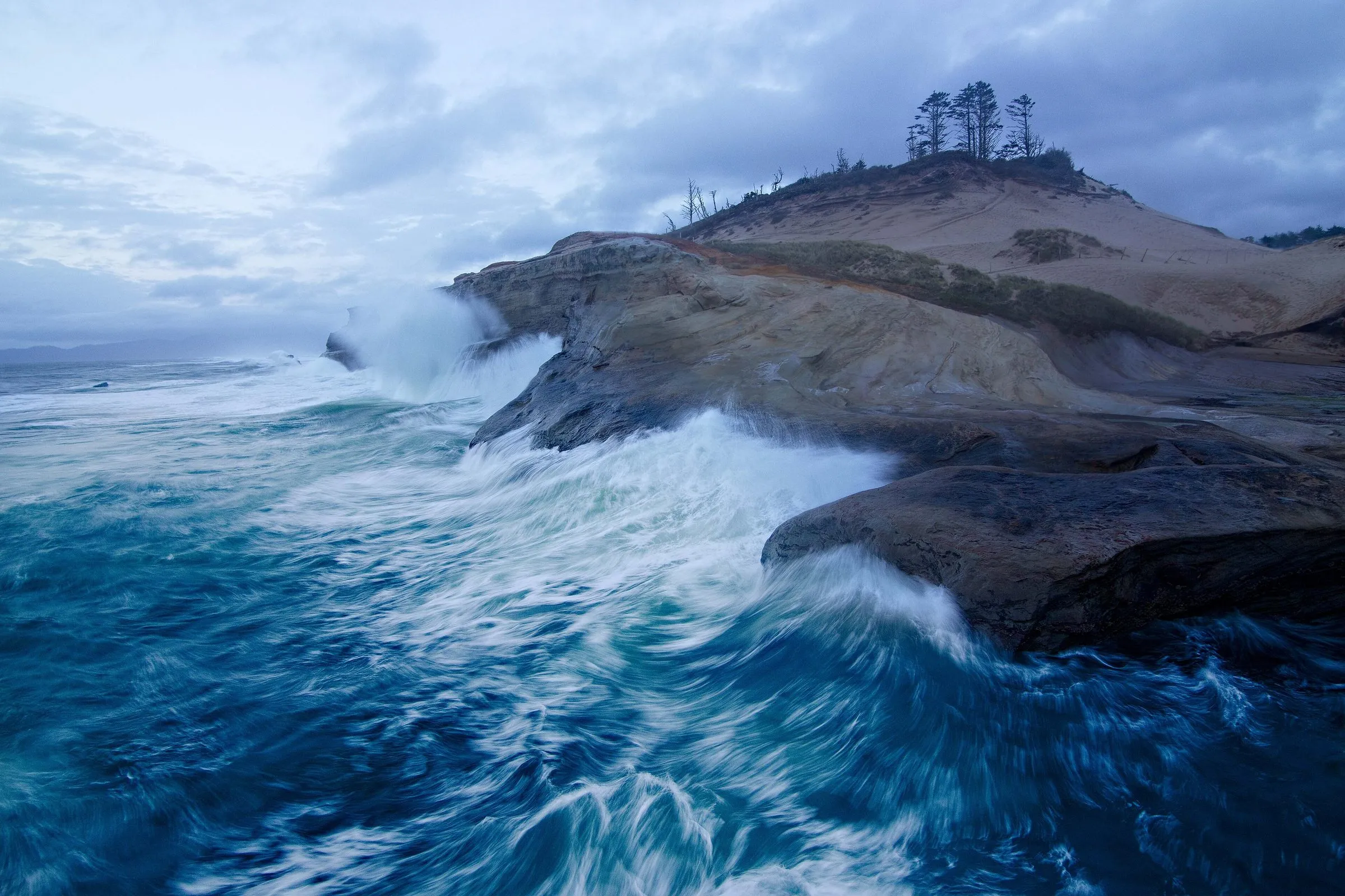 Waves breaking over the sandstone ledges of Cape Kiwanda at dusk, with a few trees on top of the headland.