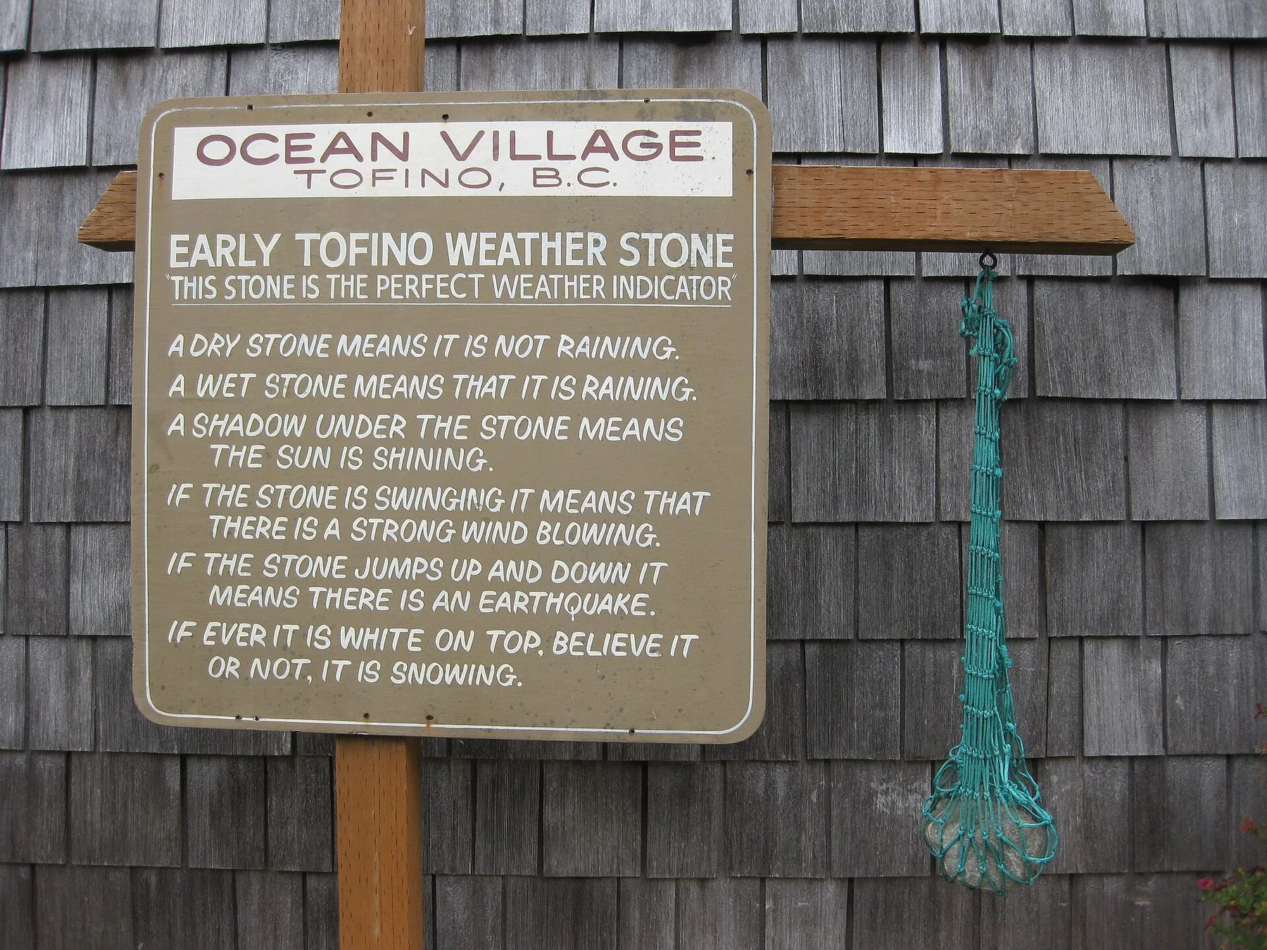 A painted sign on a shingled wall headed Early Tofino Weather Stone, with a piece of green fishing net hanging beside it.