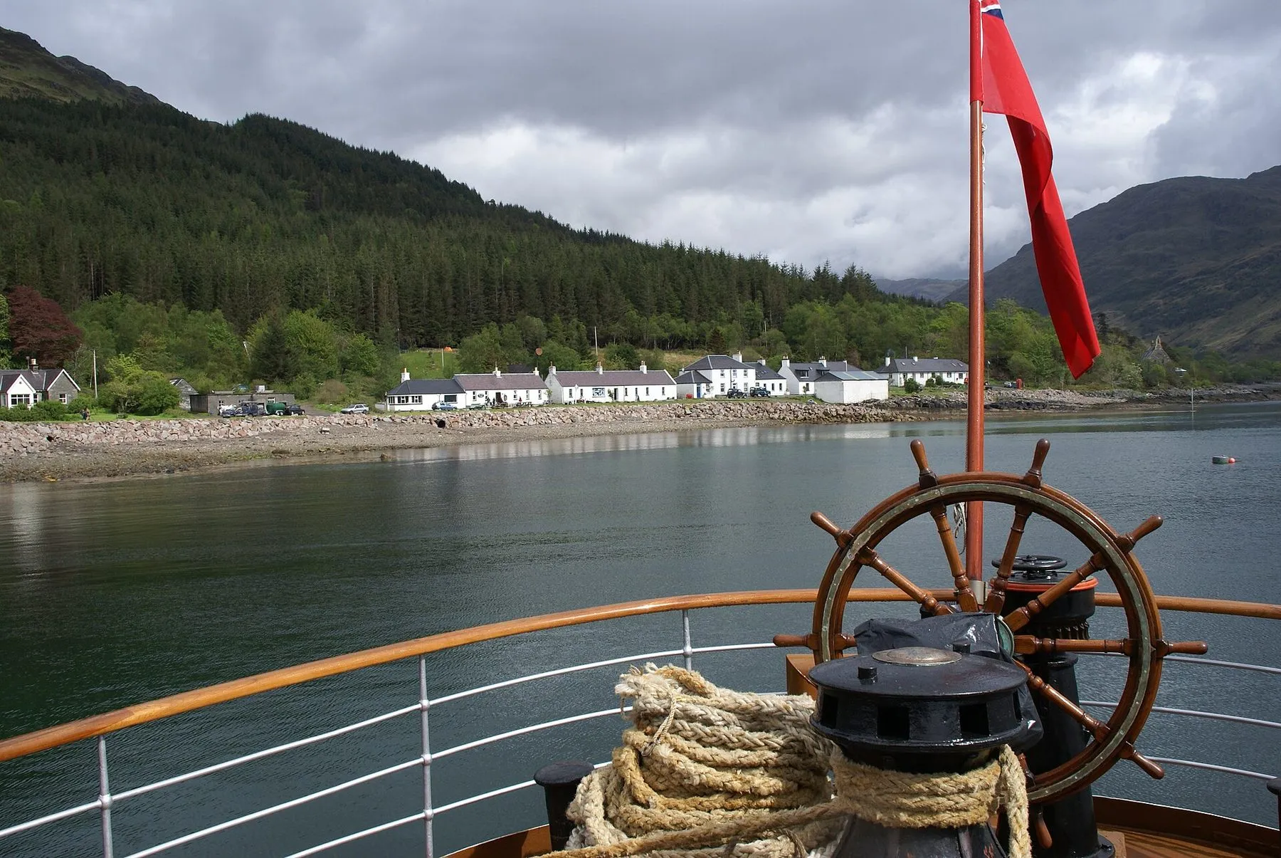 A wooden ship’s wheel, a coil of rope and a red ensign at Waverley’s stern, with a white village on a wooded shore beyond.