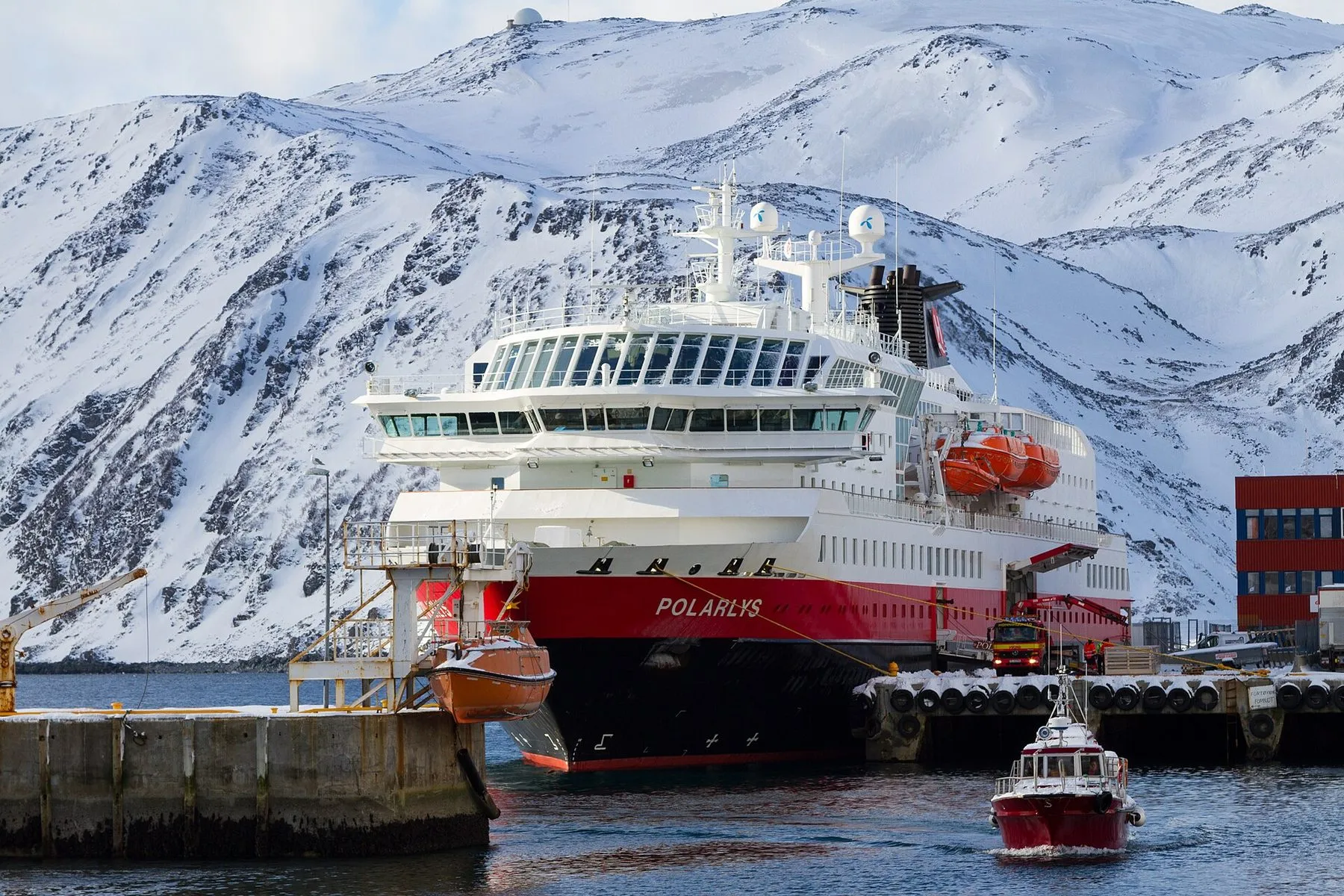 The Hurtigruten ship MS Polarlys at a quay in a small northern port, with snow-covered mountains rising behind.