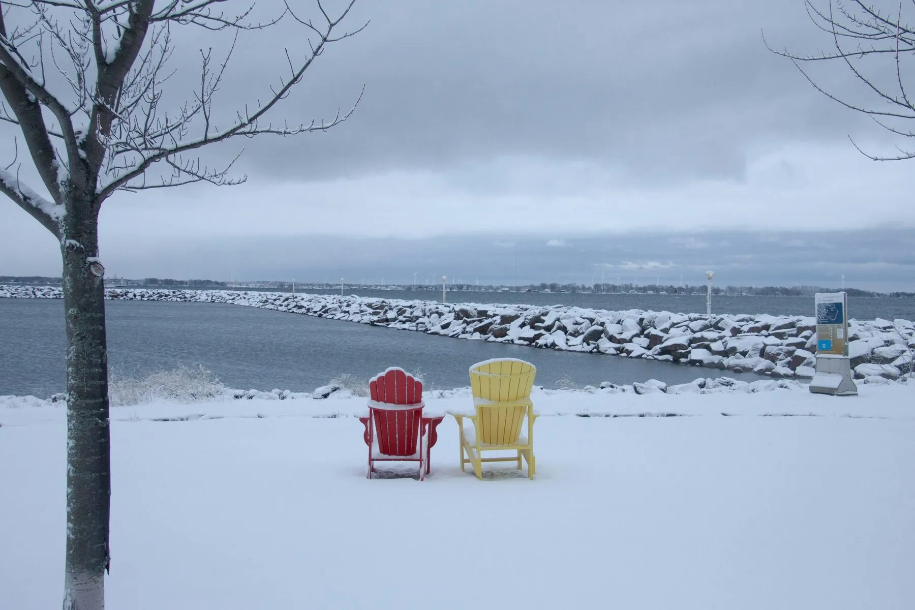 A red chair and a yellow chair side by side in deep snow beside a frozen shore, under a grey sky.
