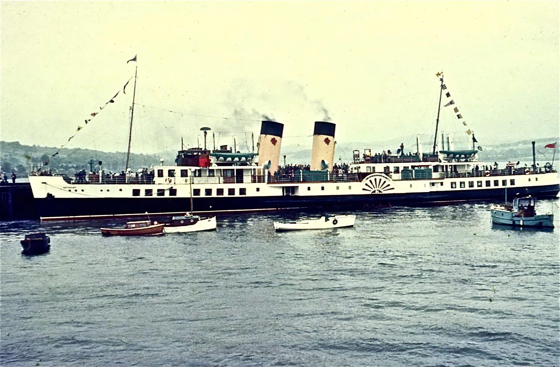 A 1970 colour photo of Waverley dressed with flags, her two funnels painted yellow with black tops, with small boats in the foreground.